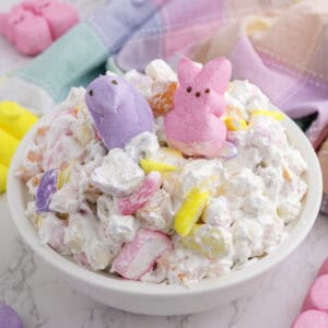 A white bowl filled with marshmallow salad, topped with pastel-colored marshmallow chick and bunny candies, with a pastel napkin in the background.