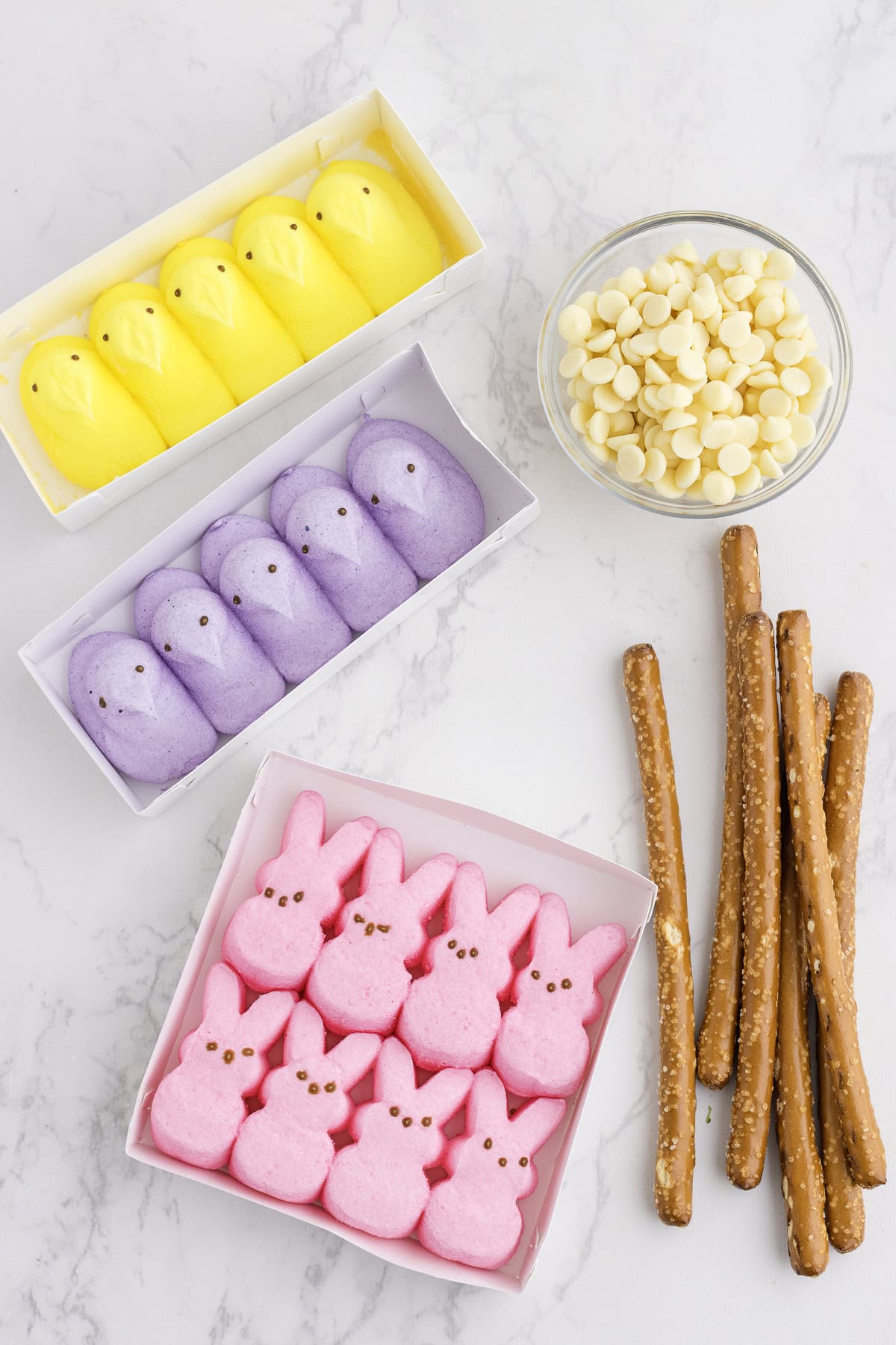 Yellow chick, purple bunny, and pink bunny marshmallow candies in boxes, white chocolate chips in a bowl, and pretzel rods on a marble surface.