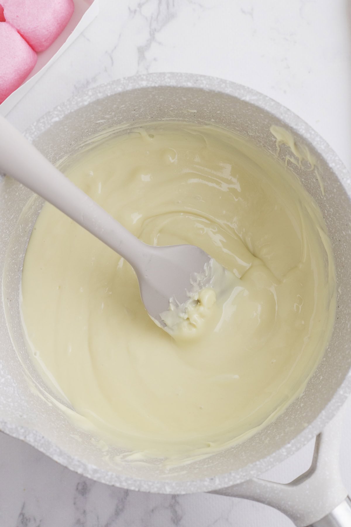 A white saucepan filled with melted white chocolate being stirred with a white spatula; pink marshmallows are visible in the upper left corner.