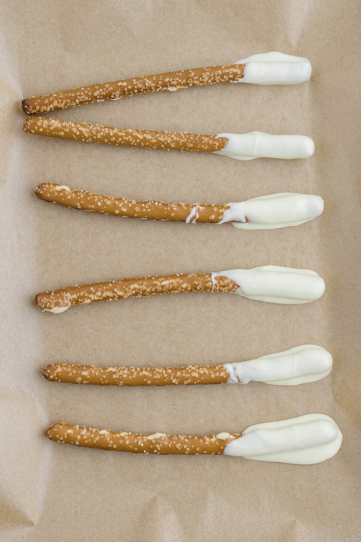 Six pretzel rods partially dipped in white chocolate are arranged in a row on brown parchment paper.