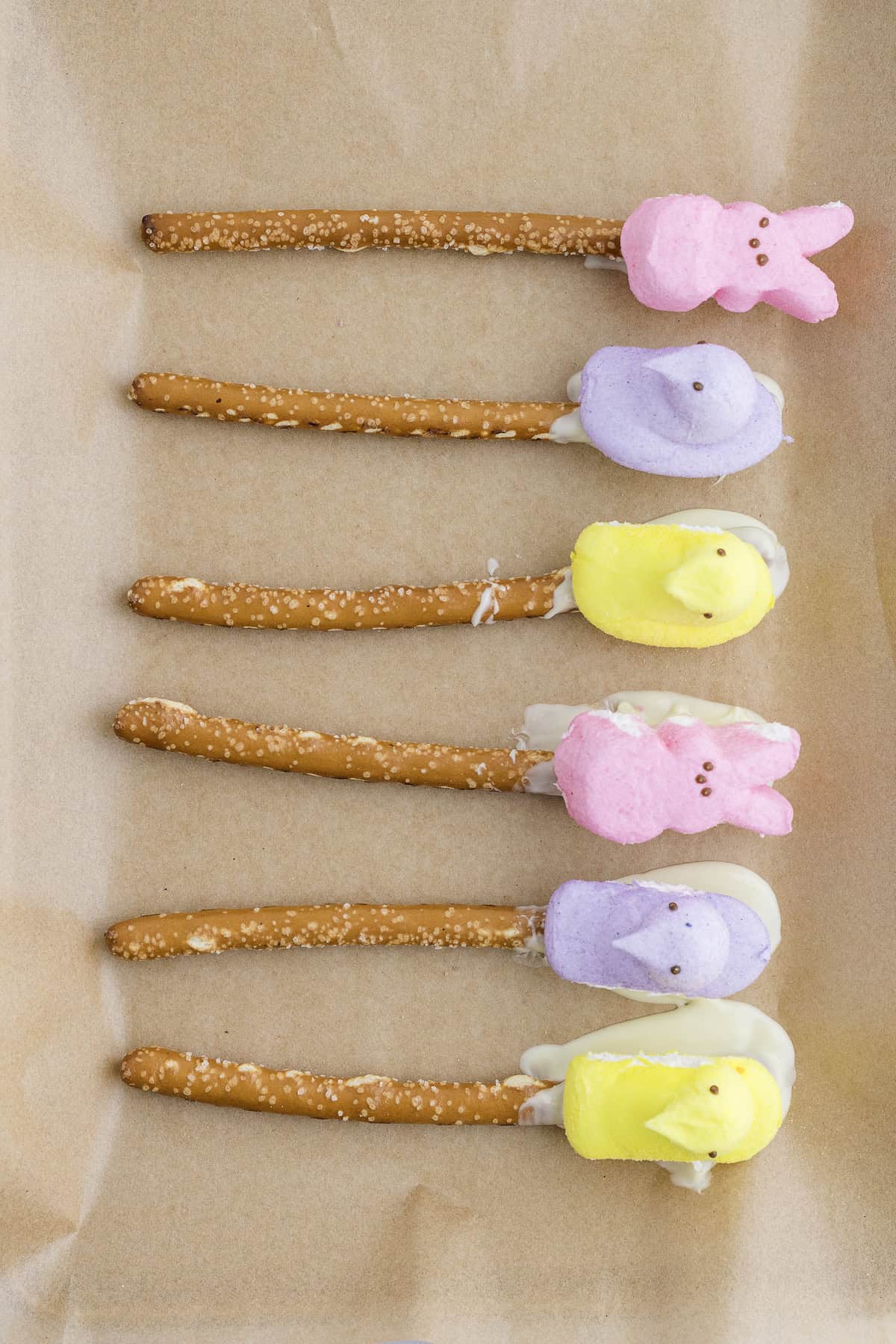 Six pretzel rods coated with white chocolate and topped with colorful marshmallow chicks and bunnies are arranged in a row on parchment paper.