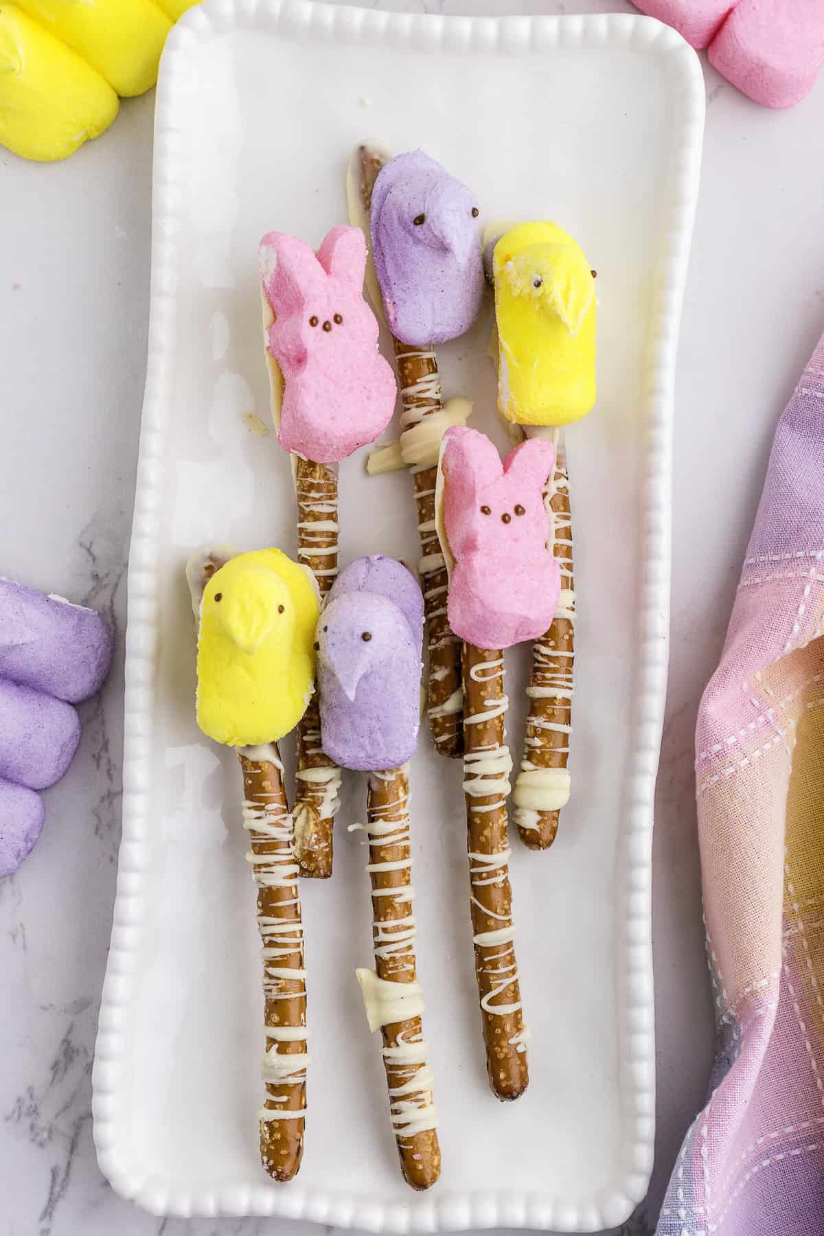 Pretzel rods drizzled with white chocolate and topped with marshmallow chicks and bunnies in yellow, pink, and purple, arranged on a white rectangular plate.