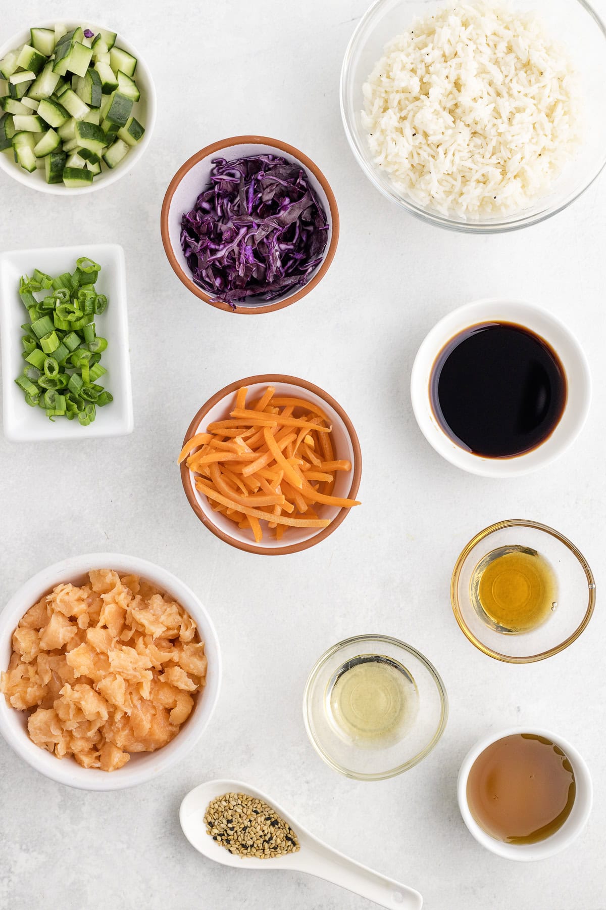 Assorted poke bowl ingredients arranged on a white surface, including diced cucumber, shredded cabbage, sliced carrots, cooked rice, green onions, raw salmon, sauces, and seasonings in bowls.