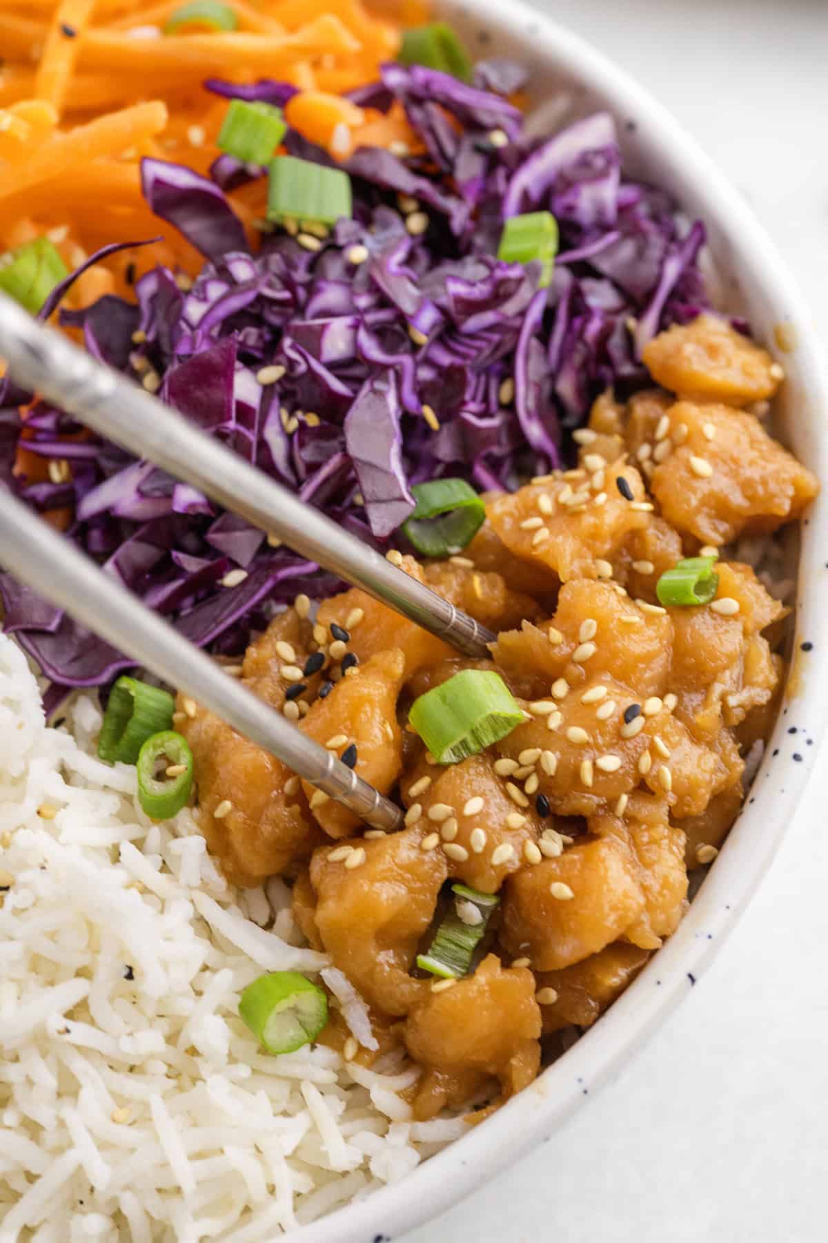 A close-up of a bowl with white rice, salmon, shredded purple cabbage, grated carrots, green onions, and sesame seeds, with chopsticks picking up chicken.