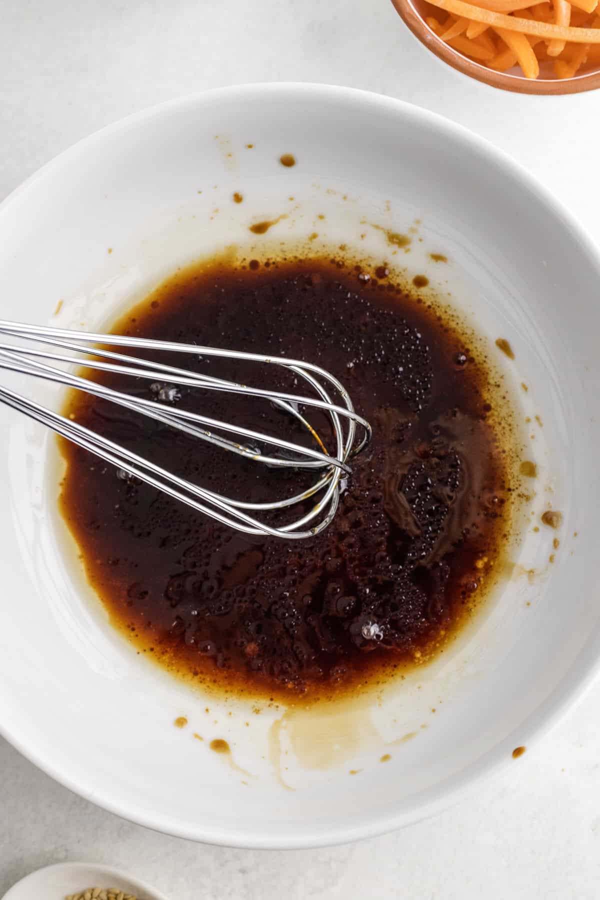 A metal whisk rests in a white bowl filled with a dark brown liquid mixture, likely a sauce or marinade, on a light surface.