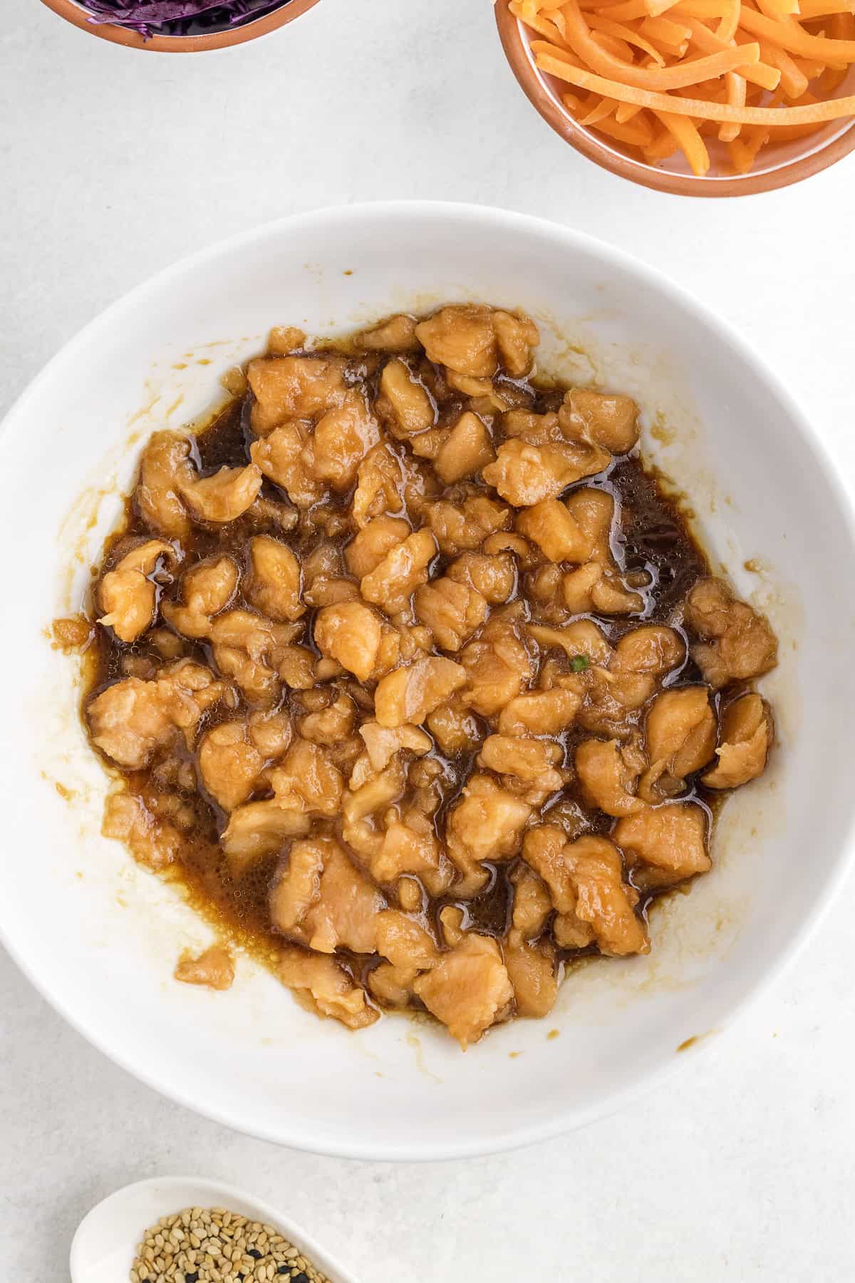 A white bowl filled with diced salmon marinating in a brown sauce sits on a white surface, surrounded by small bowls of vegetables and sesame seeds.