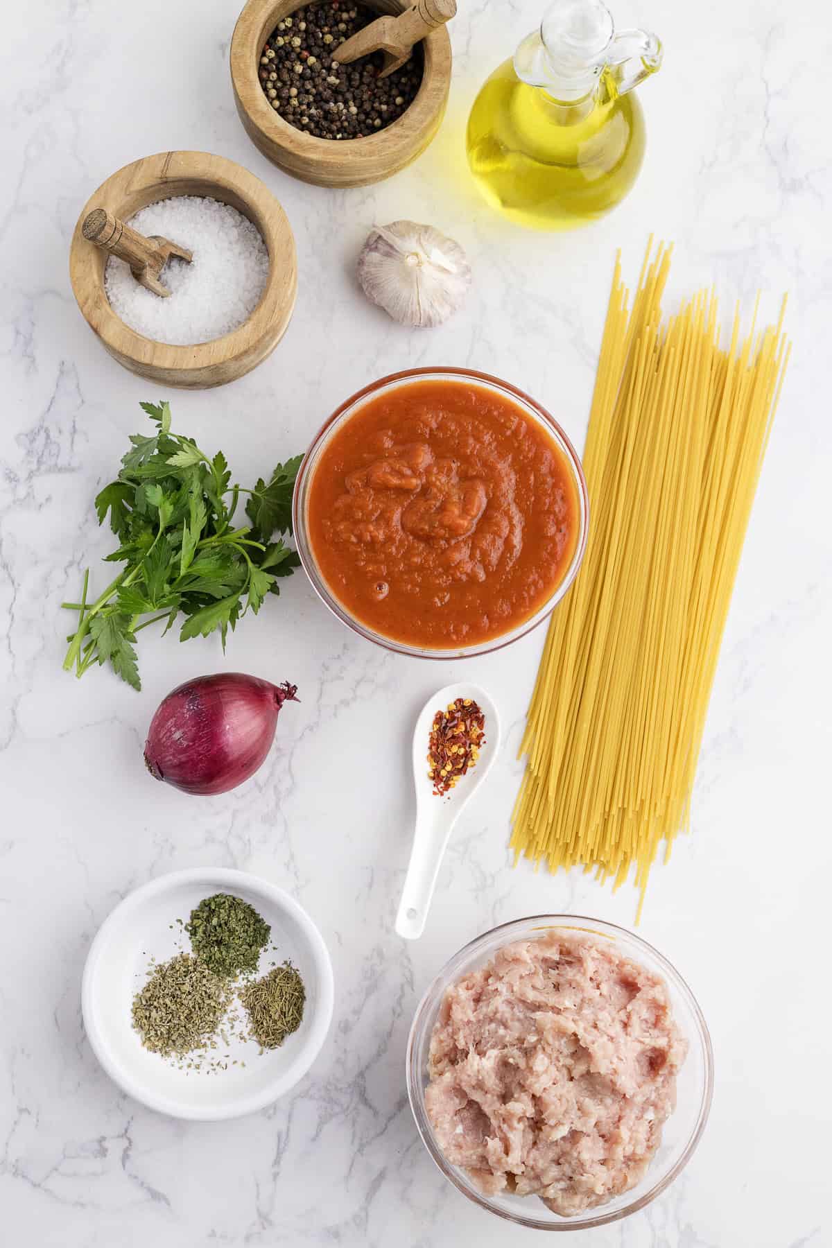 Ingredients for turkey spaghetti sauce laid out on a white surface, including dry pasta, ground turkey, tomato sauce, onion, garlic, parsley, olive oil, salt, pepper, Italian herbs, and red pepper flakes.