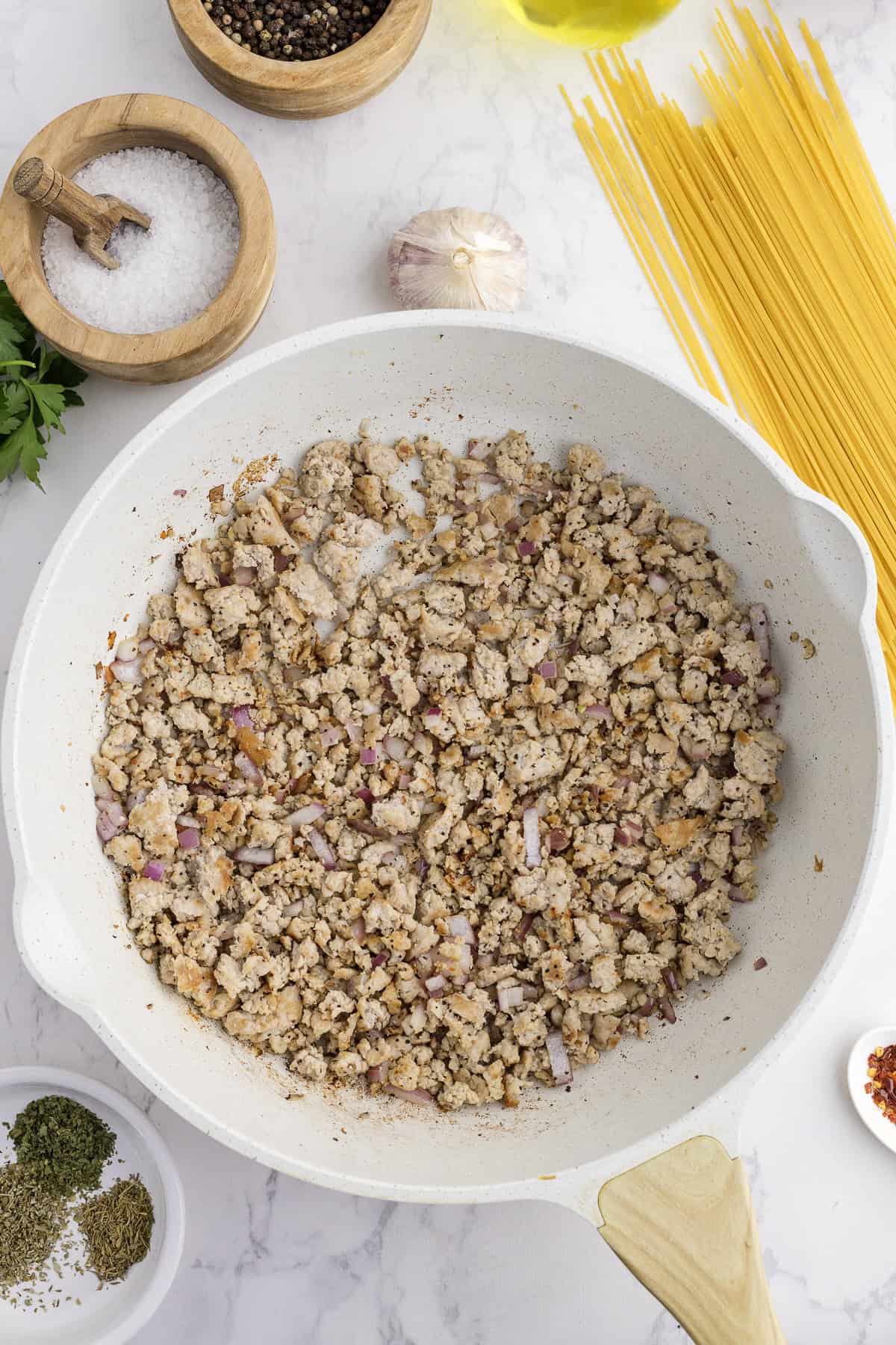 Ground turkey with chopped onions cooking in a white skillet, surrounded by uncooked spaghetti, spices, salt, pepper, garlic, and olive oil on a white countertop.