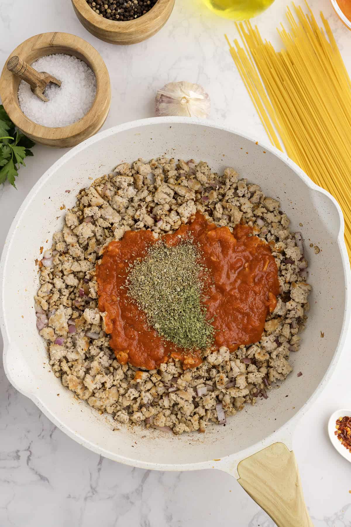 A white skillet with cooked ground turkey, tomato sauce, and herbs sits on a marble counter surrounded by salt, pepper, garlic, parsley, dry spaghetti, and olive oil.