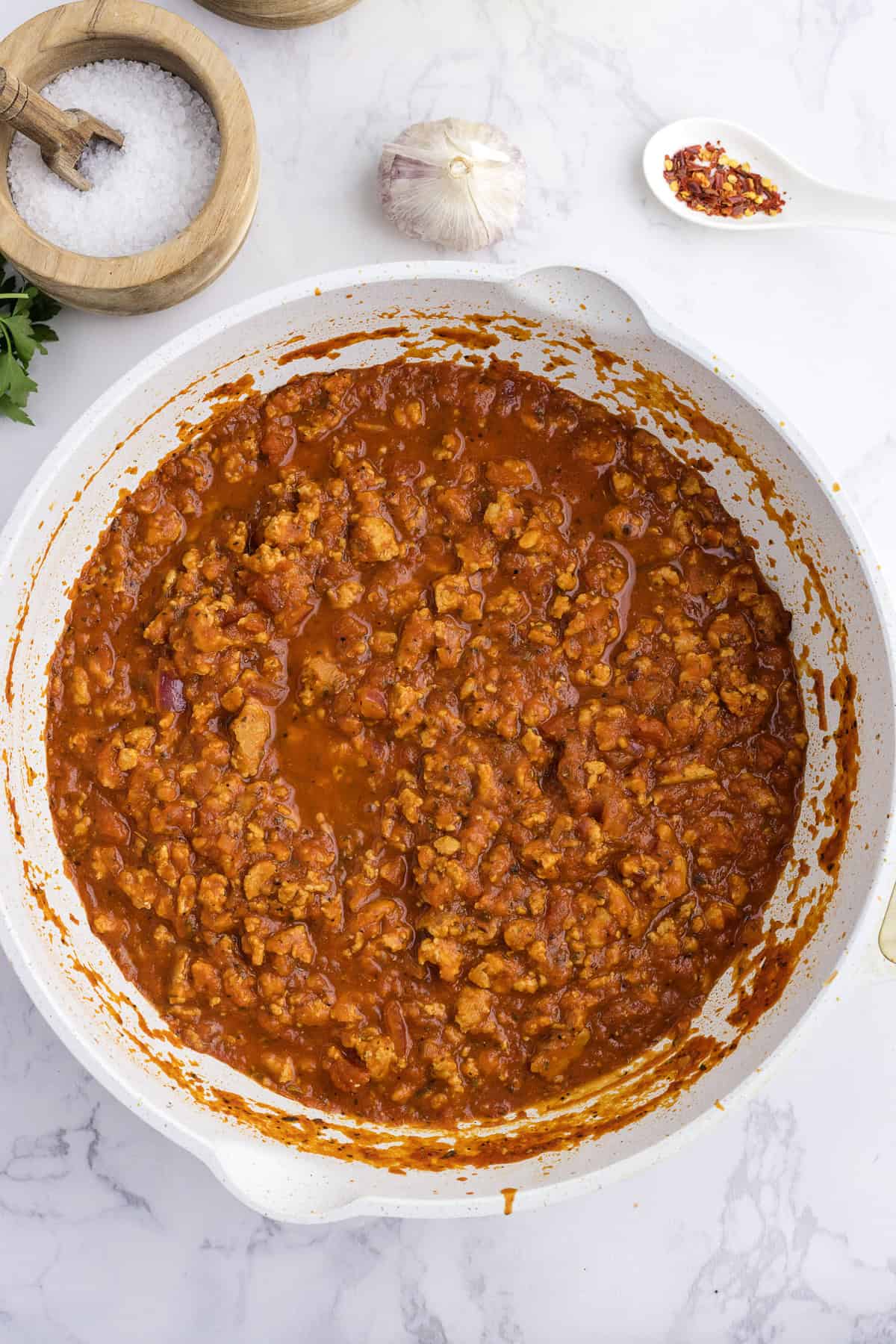 A white skillet filled with cooked Turkey spaghetti sauce sits on a marble surface, with a bowl of salt, a garlic bulb, and a spoon of red pepper flakes nearby.
