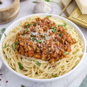 A bowl of turkey spaghetti topped with meat sauce, grated cheese, and fresh parsley, with garlic and a cheese grater in the background.