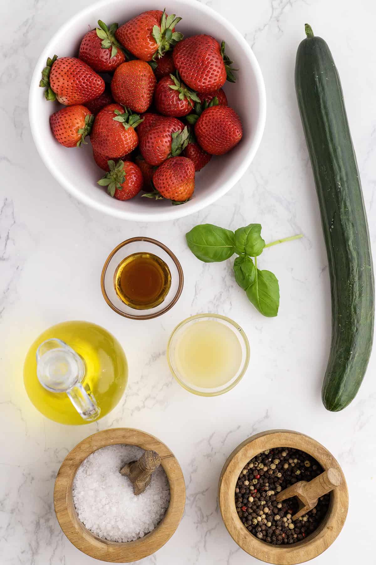 A bowl of strawberries, a whole cucumber, basil leaves, a glass bottle of olive oil, a small jar of vinegar, and wooden bowls of salt and pepper on a marble surface.
