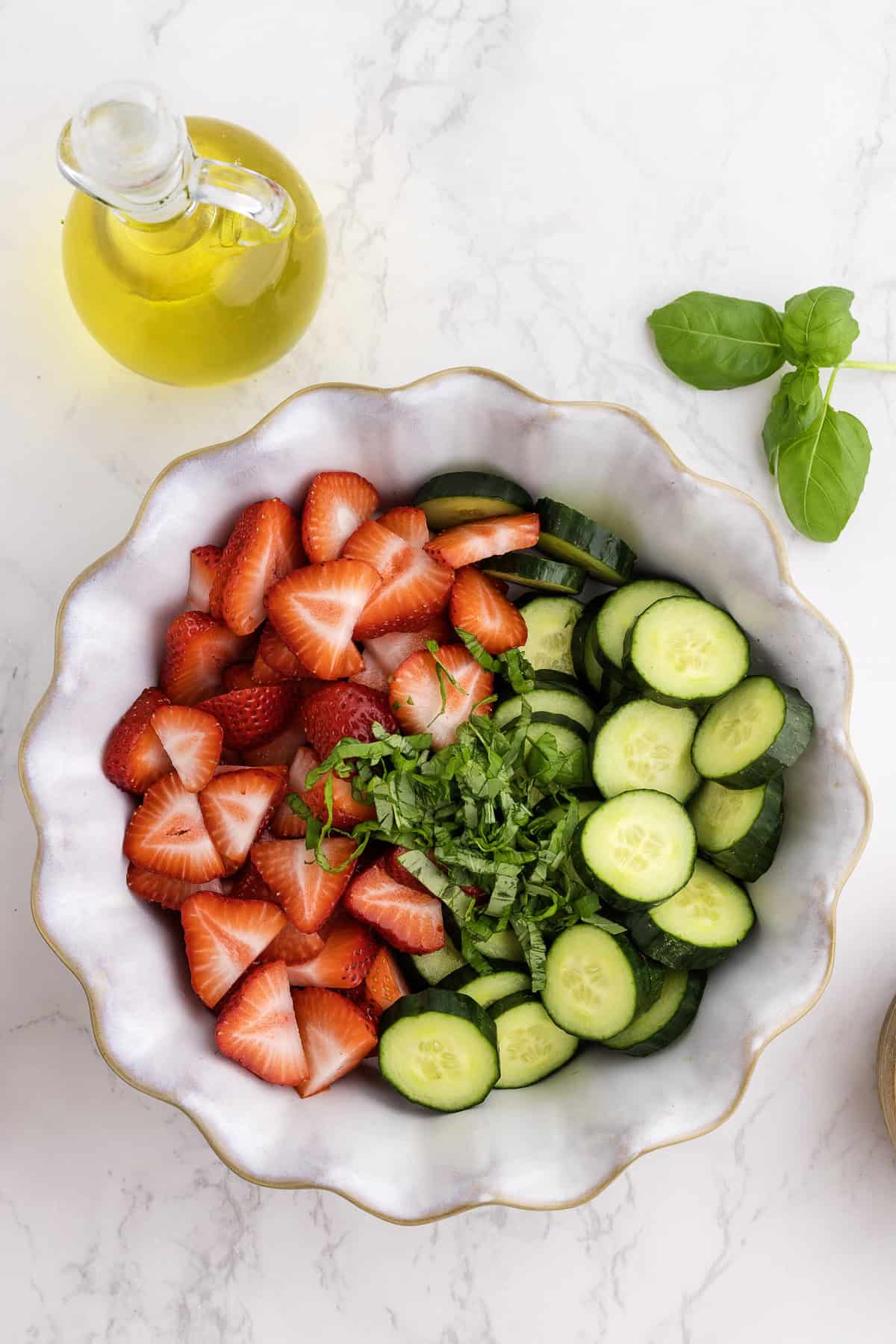 A bowl with sliced strawberries, cucumbers, and chopped basil on a marble surface, next to a glass jug of olive oil and a sprig of fresh basil.