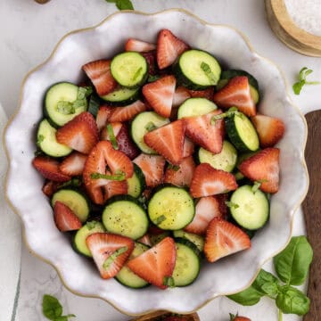 A bowl filled with sliced cucumbers and strawberries, garnished with fresh herbs, sits on a white surface.