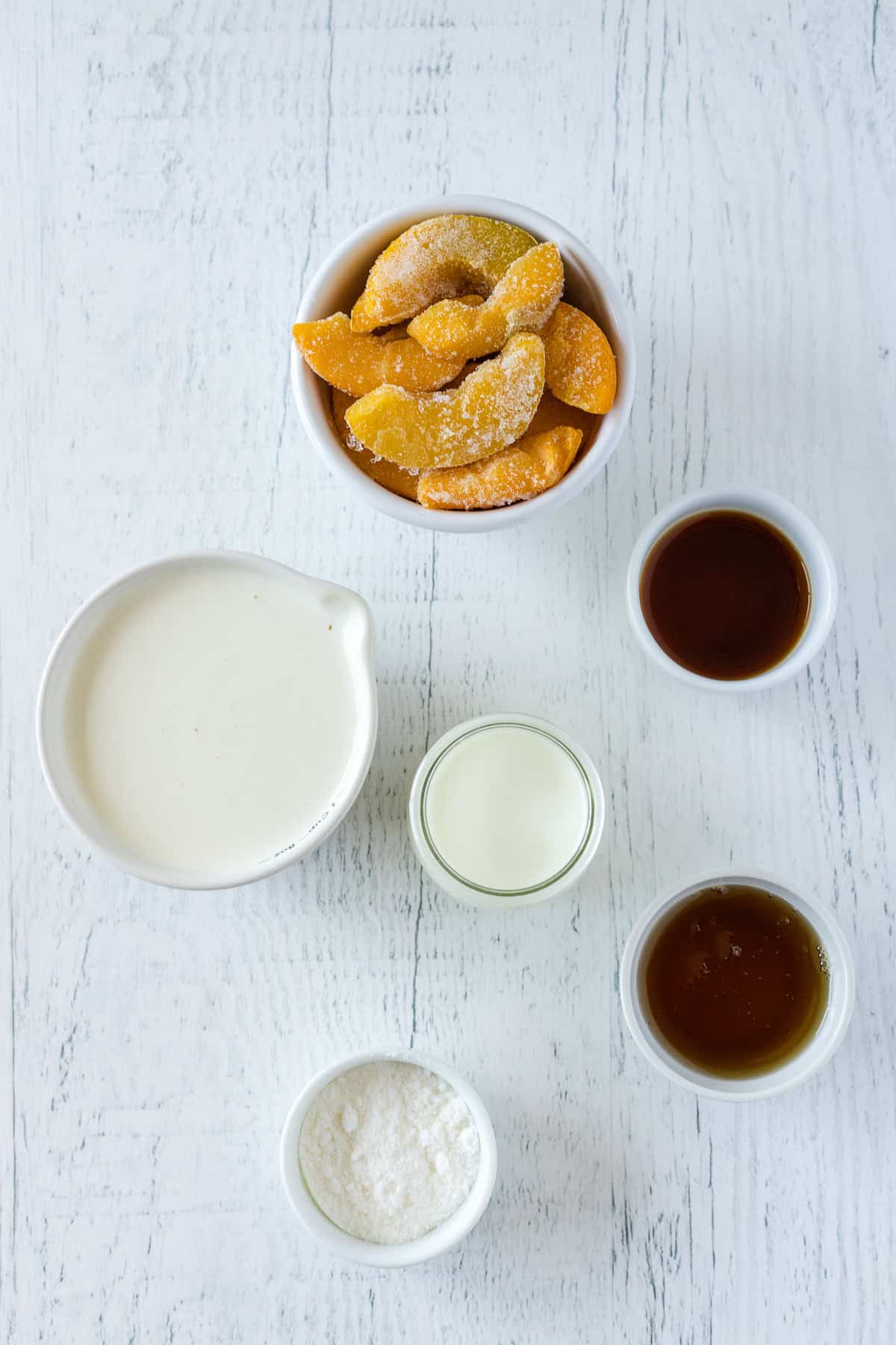 Five small bowls containing frozen peach slices, heavy cream, milk, vanilla pudding, vanilla extract, and maple syrup, arranged on a white wooden surface.