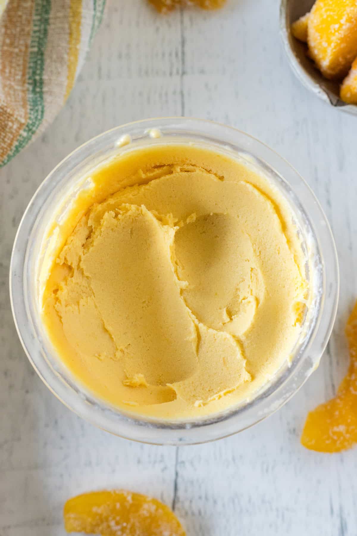 A glass bowl filled with smooth, orange-colored ice cream is shown from above, with pieces of orange fruit and a striped towel nearby on a white surface.