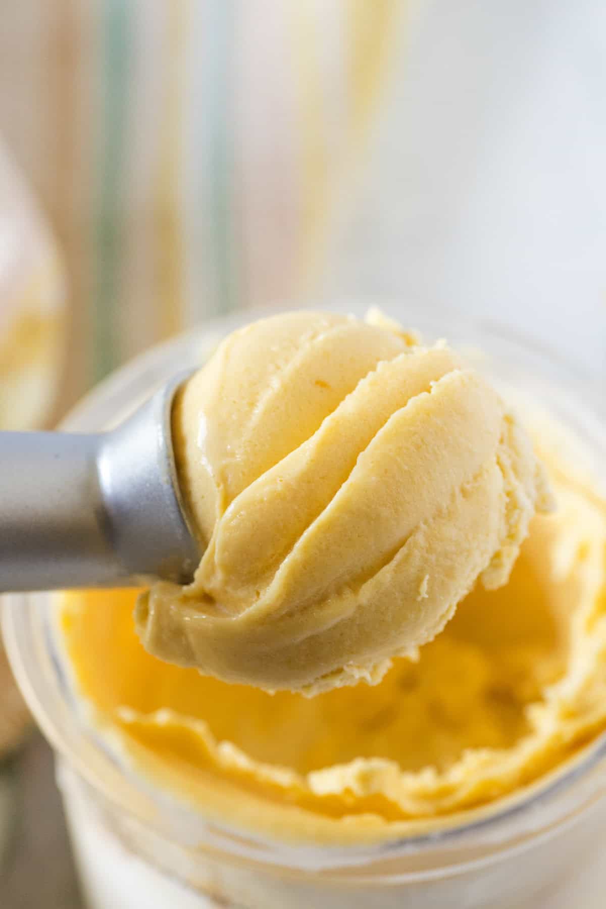 A close-up of a metal ice cream scoop lifting a scoop of peach ice cream from a container.