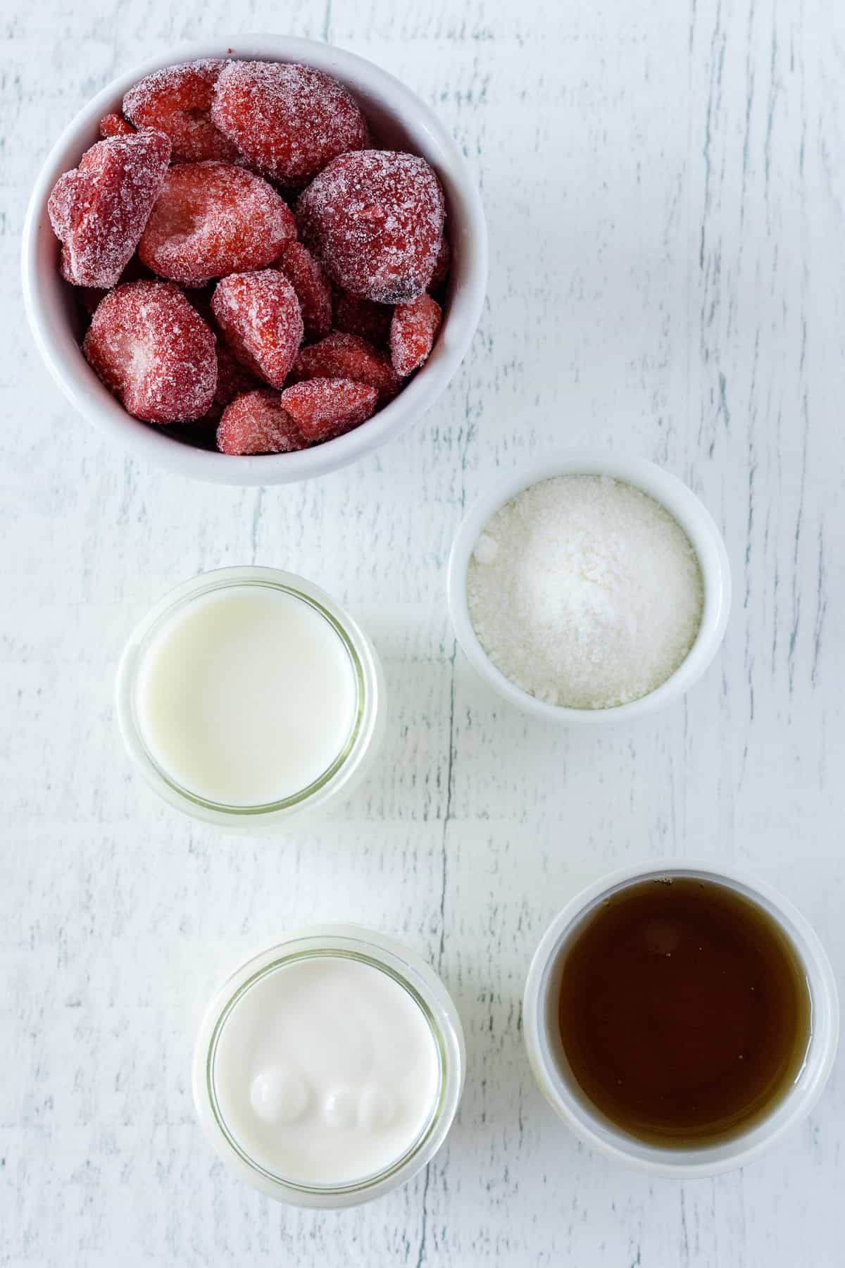 Five small bowls containing frozen strawberries, granulated sugar, milk, cream, and a brown liquid, arranged on a white wooden surface.