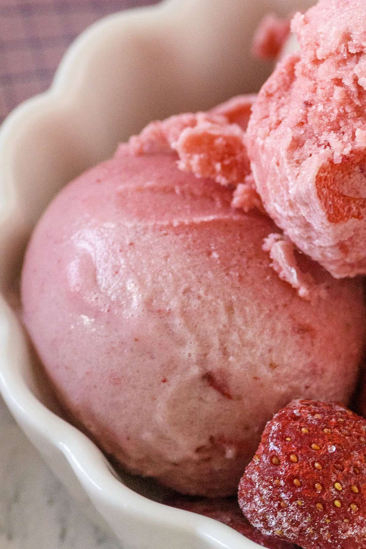 Two scoops of pink strawberry ice cream in a white bowl, with a whole strawberry placed in front.