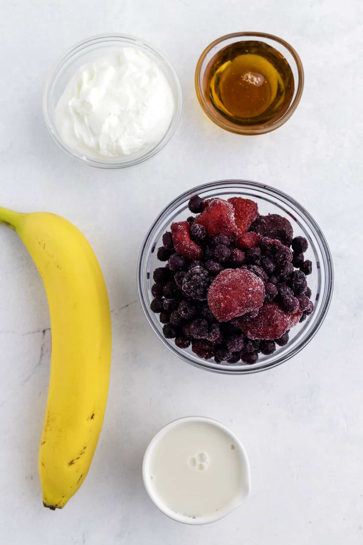 A banana, a bowl of frozen mixed berries, a bowl of yogurt, a small bowl of maple syrup, and a small cup of milk are arranged on a white surface.