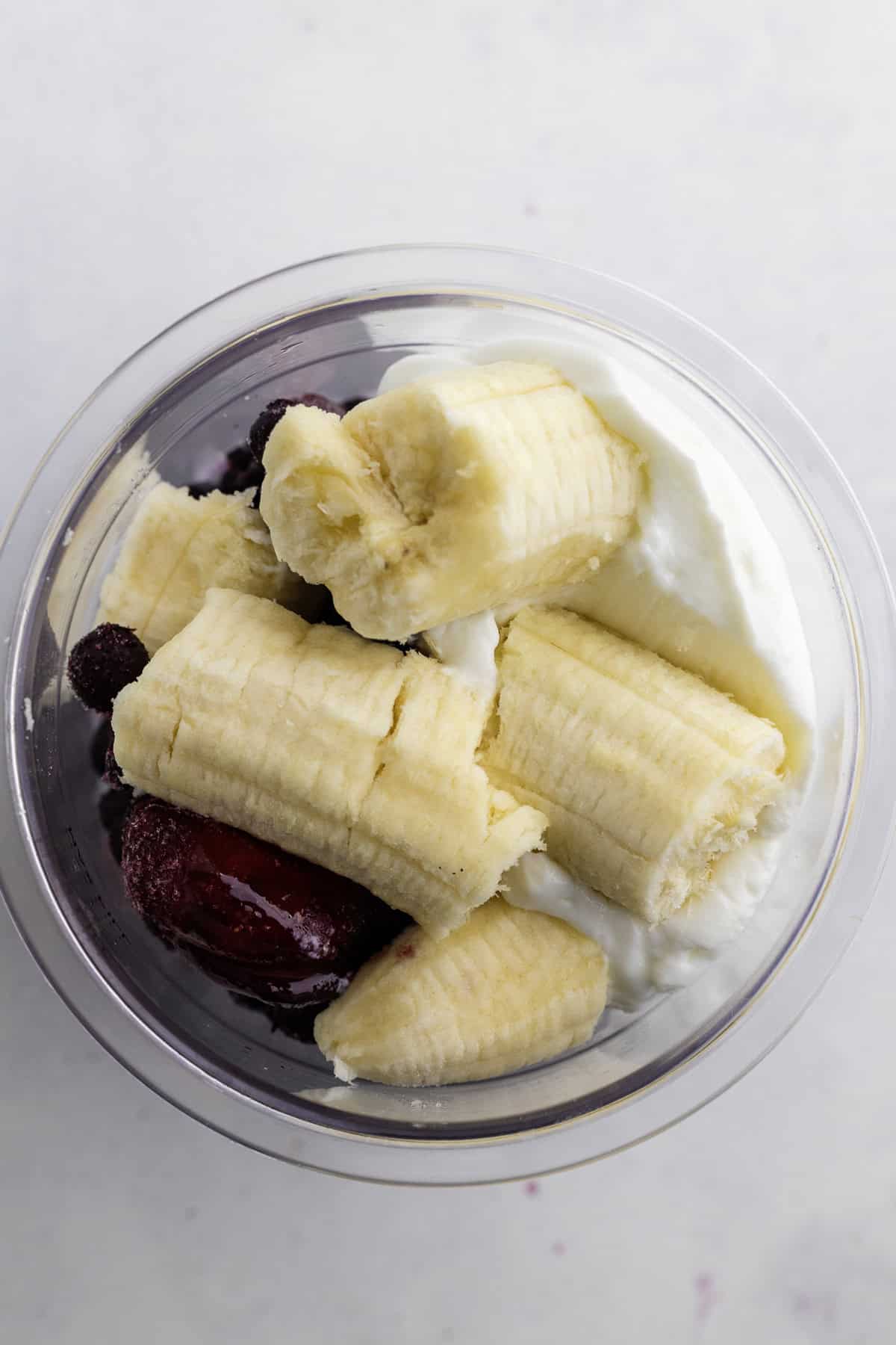 A clear bowl containing sliced banana, mixed berries, and yogurt, viewed from above on a white surface.