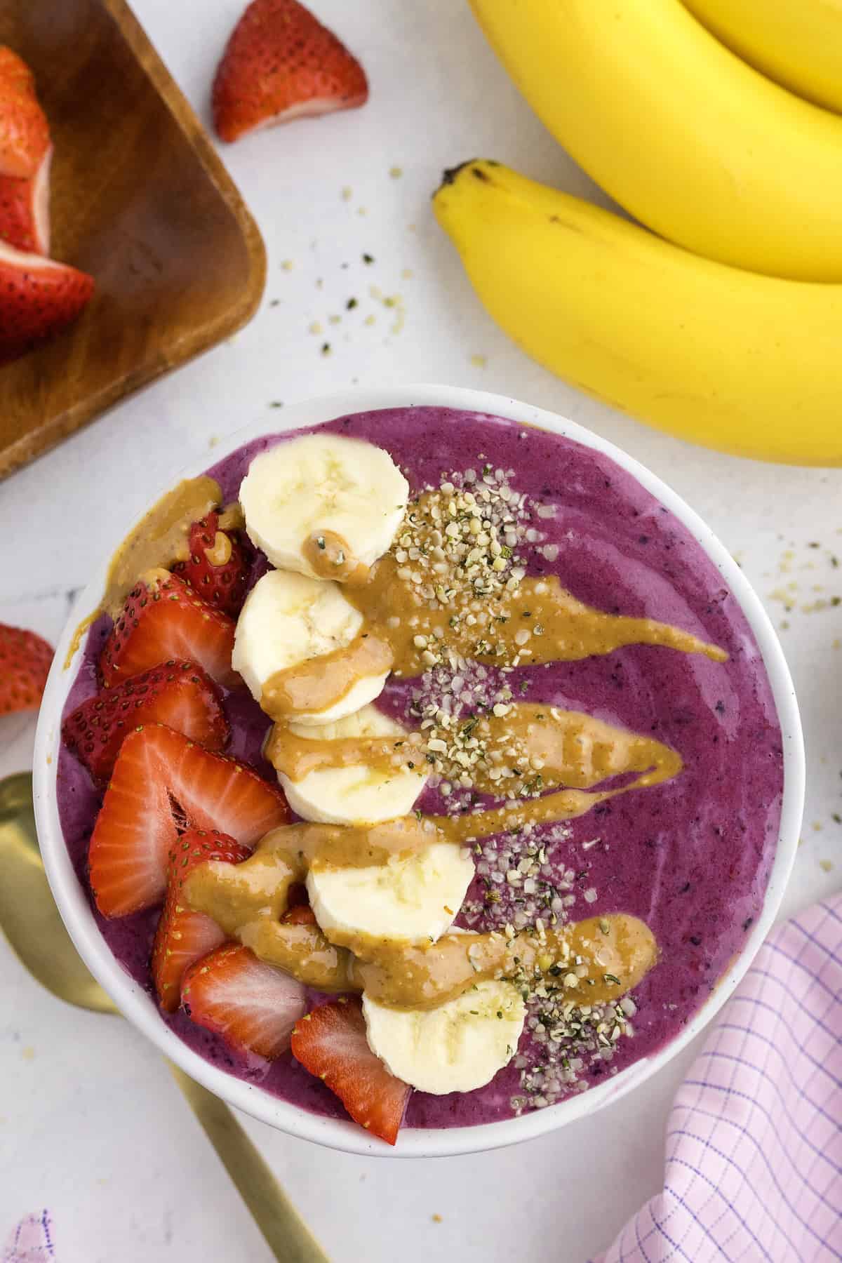 A smoothie bowl topped with banana slices, strawberry slices, hemp seeds, and a drizzle of nut butter, with bananas and strawberries nearby.