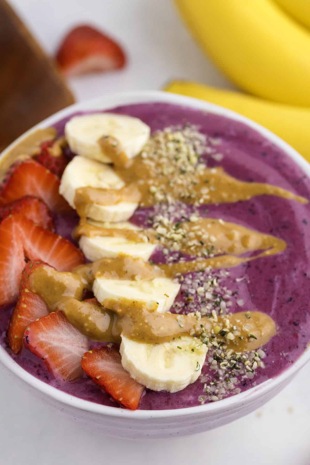 A Ninja Creami smoothie bowl topped with sliced strawberries, banana, hemp seeds, and peanut butter, with bananas and a strawberry in the background.