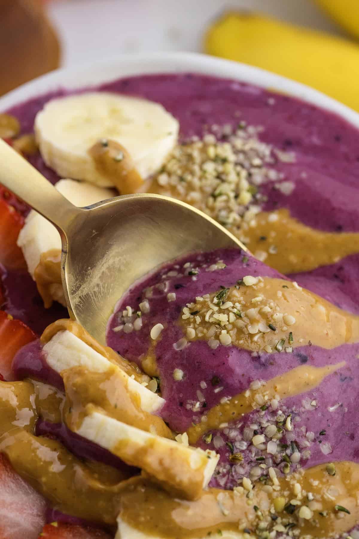 A close-up of a gold spoon in a smoothie bowl topped with banana slices, strawberries, peanut butter, and sprinkled seeds.