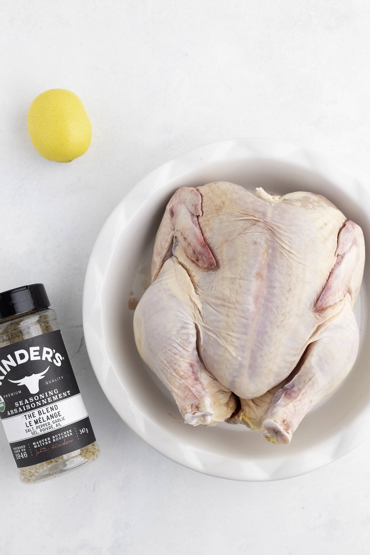Raw whole chicken in a white bowl, a bottle of seasoning blend, and a whole lemon on a white surface.