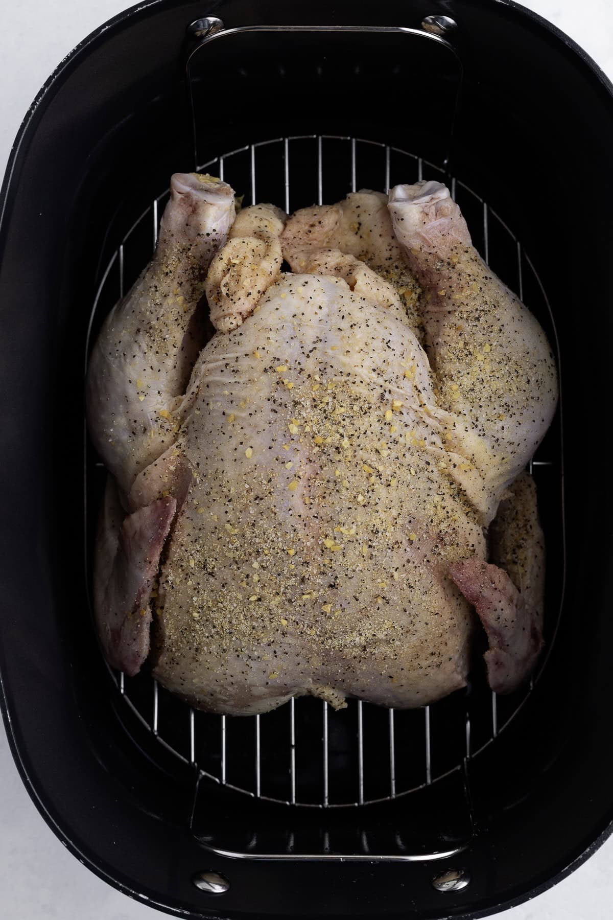 A whole raw chicken seasoned with spices sits on a rack inside a black roasting pan.