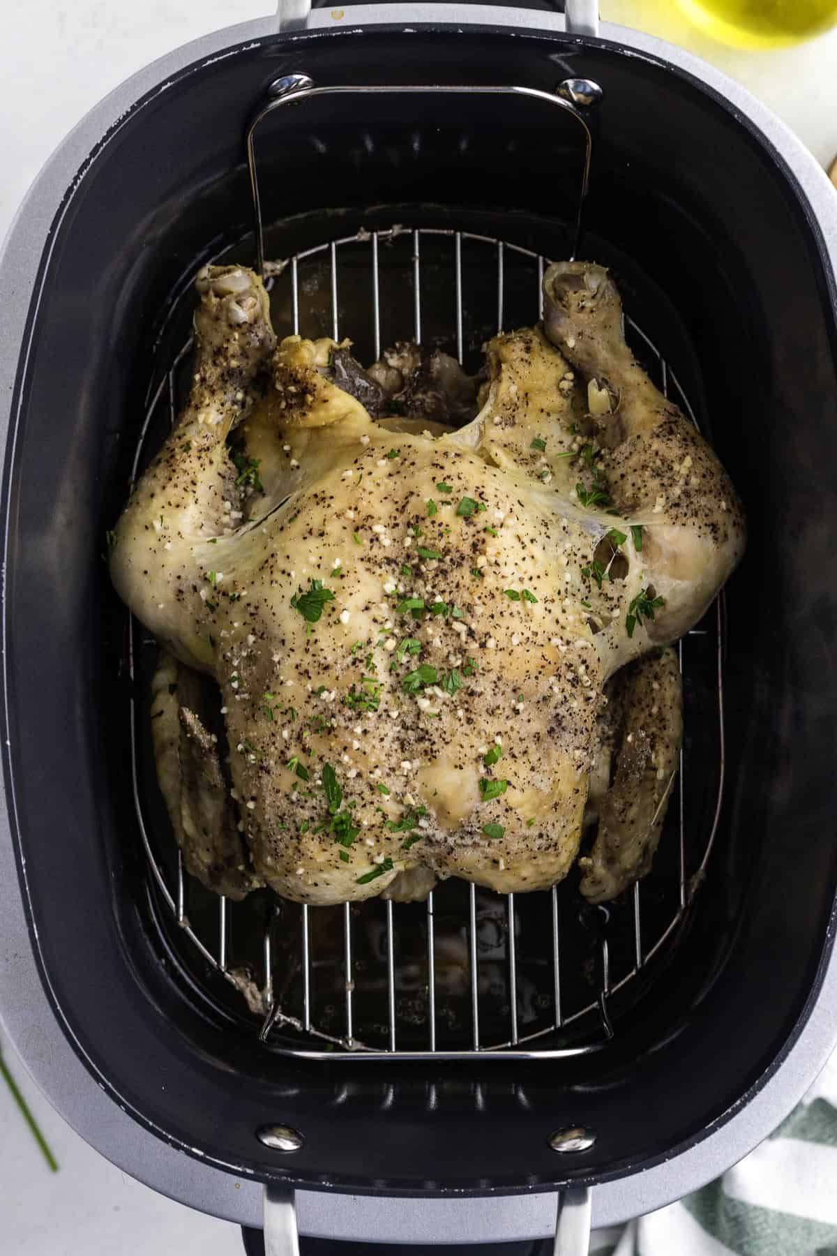 Whole seasoned chicken sits on a wire rack inside a black roasting pan, garnished with herbs and surrounded by cooking juices.