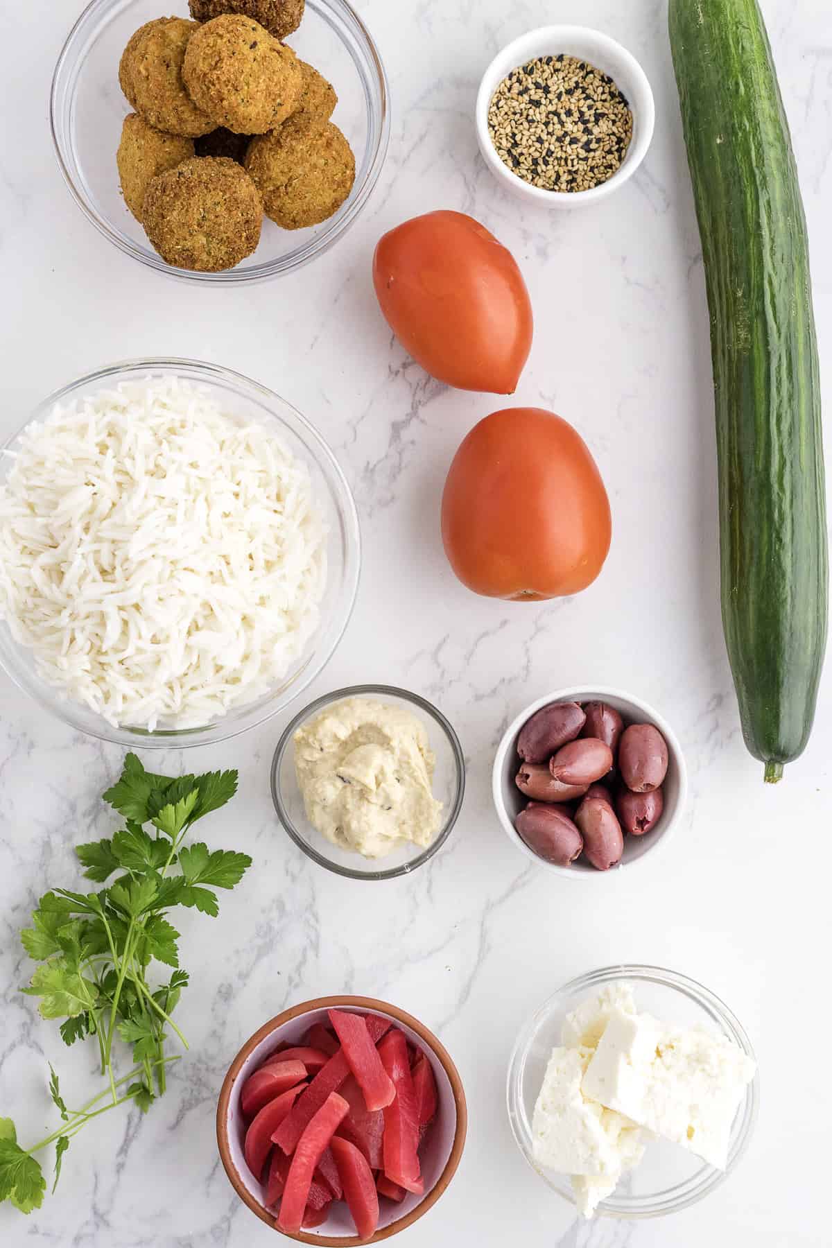 Bowls of rice, falafel, feta, hummus, olives, pickled turnip, and sesame seeds with cucumber, tomatoes, and parsley arranged on a white marble surface.