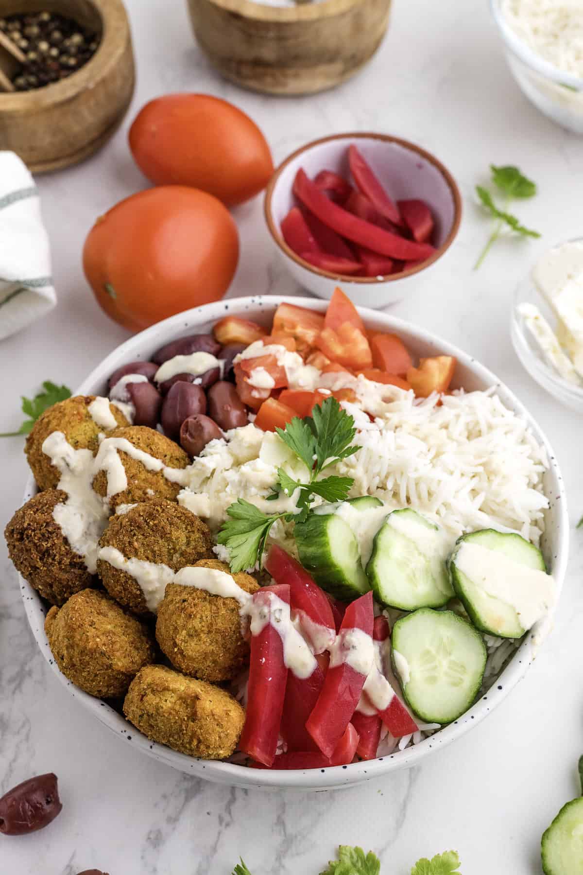 A vibrant falafel rice bowl featuring rice, sliced cucumbers, tomatoes, pickled vegetables, olives, parsley, feta cheese, and creamy tahini sauce, with whole tomatoes and fresh ingredients in the background.
