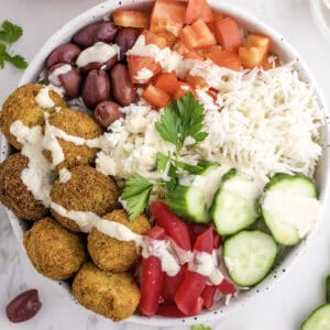 A bowl with falafel, rice, kidney beans, diced tomatoes, cucumber slices, pickled vegetables, feta cheese, kalamata olives, and tahini sauce, garnished with parsley.