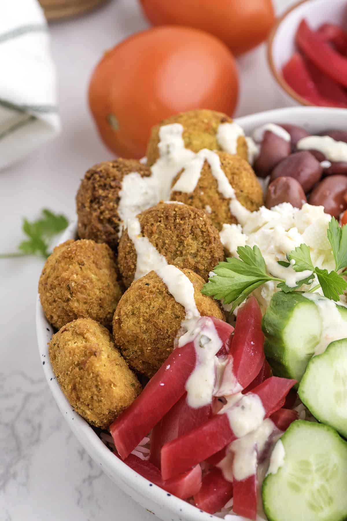 A bowl with falafel, sliced cucumbers, pickled turnips, olives, feta cheese, and parsley, topped with a drizzle of white sauce. Tomatoes are in the background.