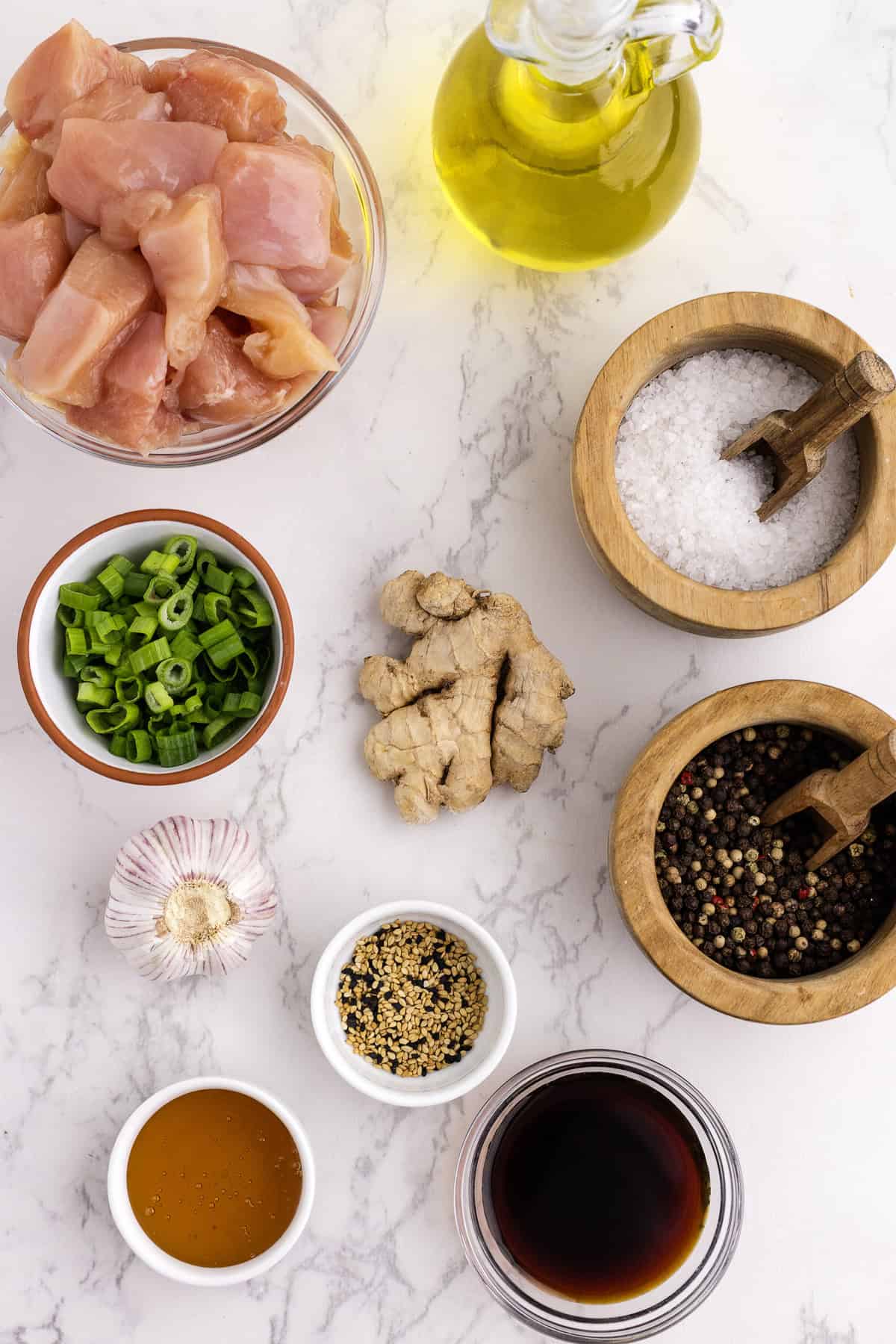 Poulet cru en dés, huile d'olive, sel, poivre, oignons verts, gingembre, ail, graines de sésame, miel et sauce soja disposés sur un plan de travail en marbre.