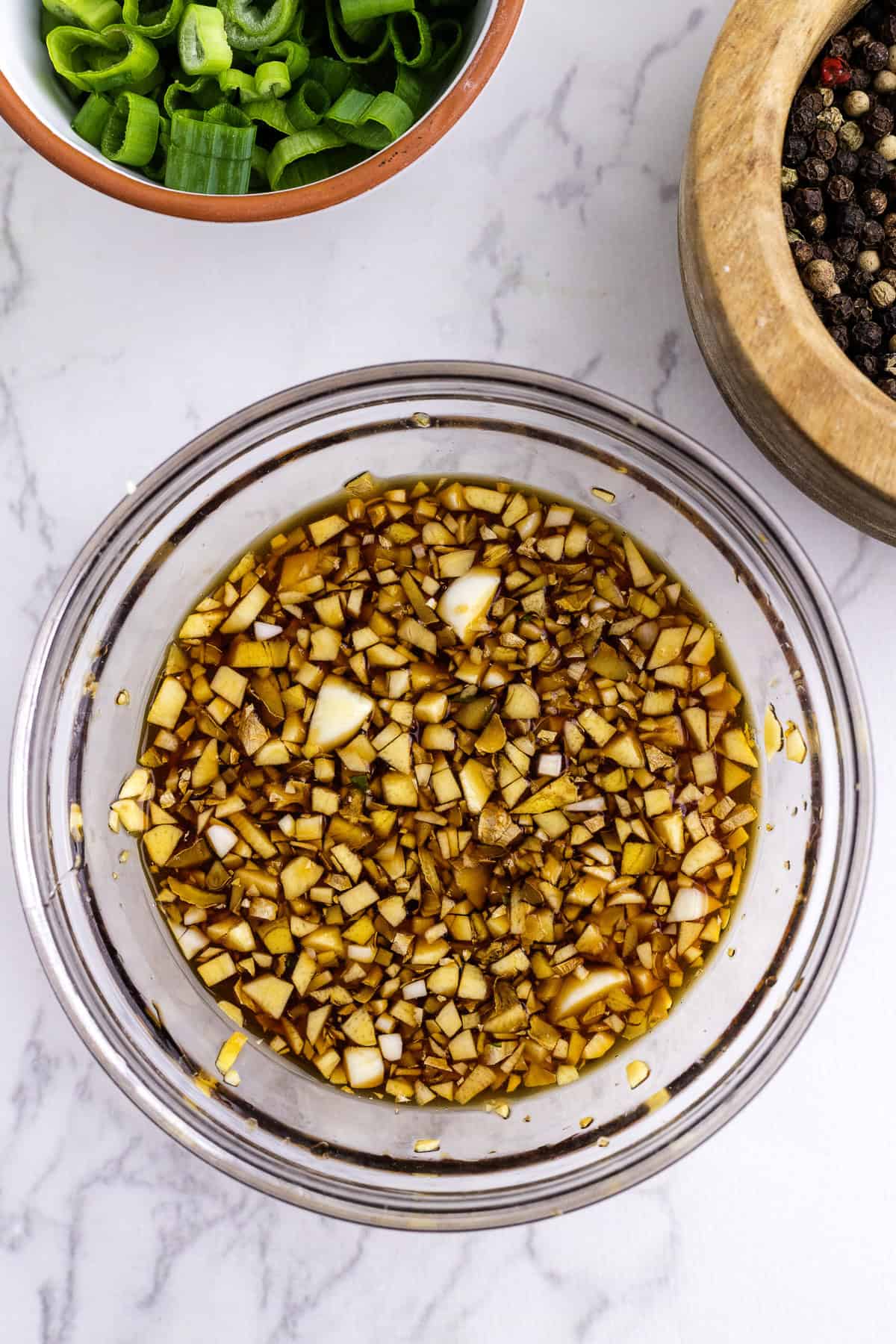 Un bol en verre rempli d'une marinade à base de soja contenant de l'ail et des oignons finement hachés, placé sur une surface en marbre près de bols d'oignons verts et de grains de poivre entiers.