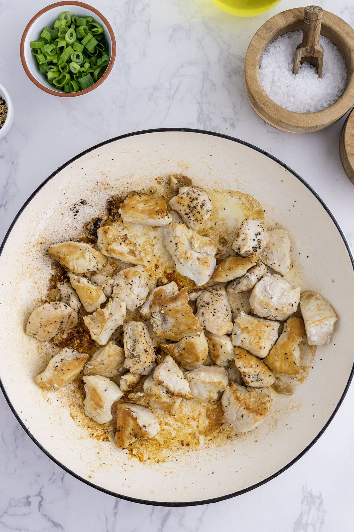 Des morceaux de blanc de poulet cuit et assaisonné dans une poêle blanche, entourés de bols d'oignons verts émincés, de sel, de poivre et d'huile d'olive sur un comptoir en marbre.