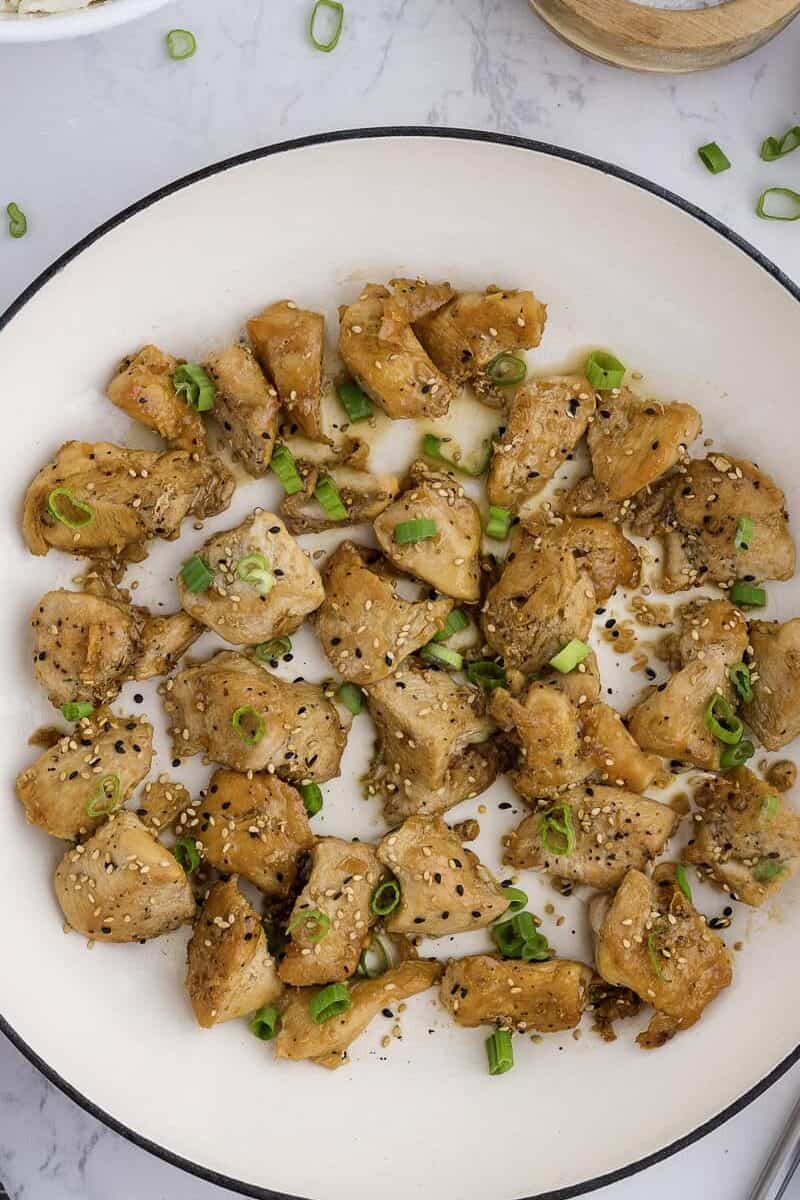 A white skillet with cooked, seasoned chicken pieces garnished with chopped green onions, surrounded by bowls of rice, salt, pepper, olive oil, and garlic on a marble countertop.