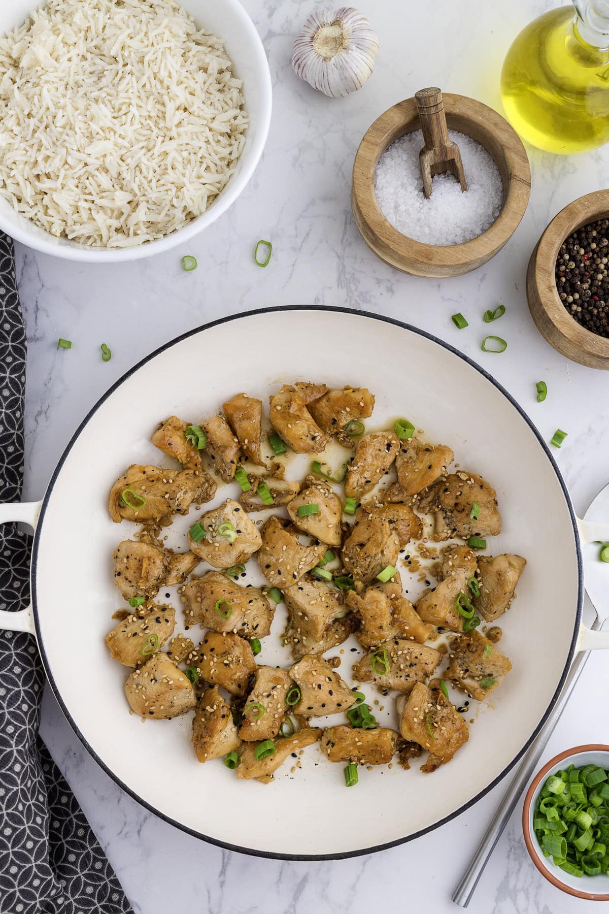 Morceaux de poulet cuits et assaisonnés dans une poêle blanche, garnis d'oignons verts, avec des bols de riz, du sel, du poivre, de l'huile et de l'ail sur un comptoir en marbre.