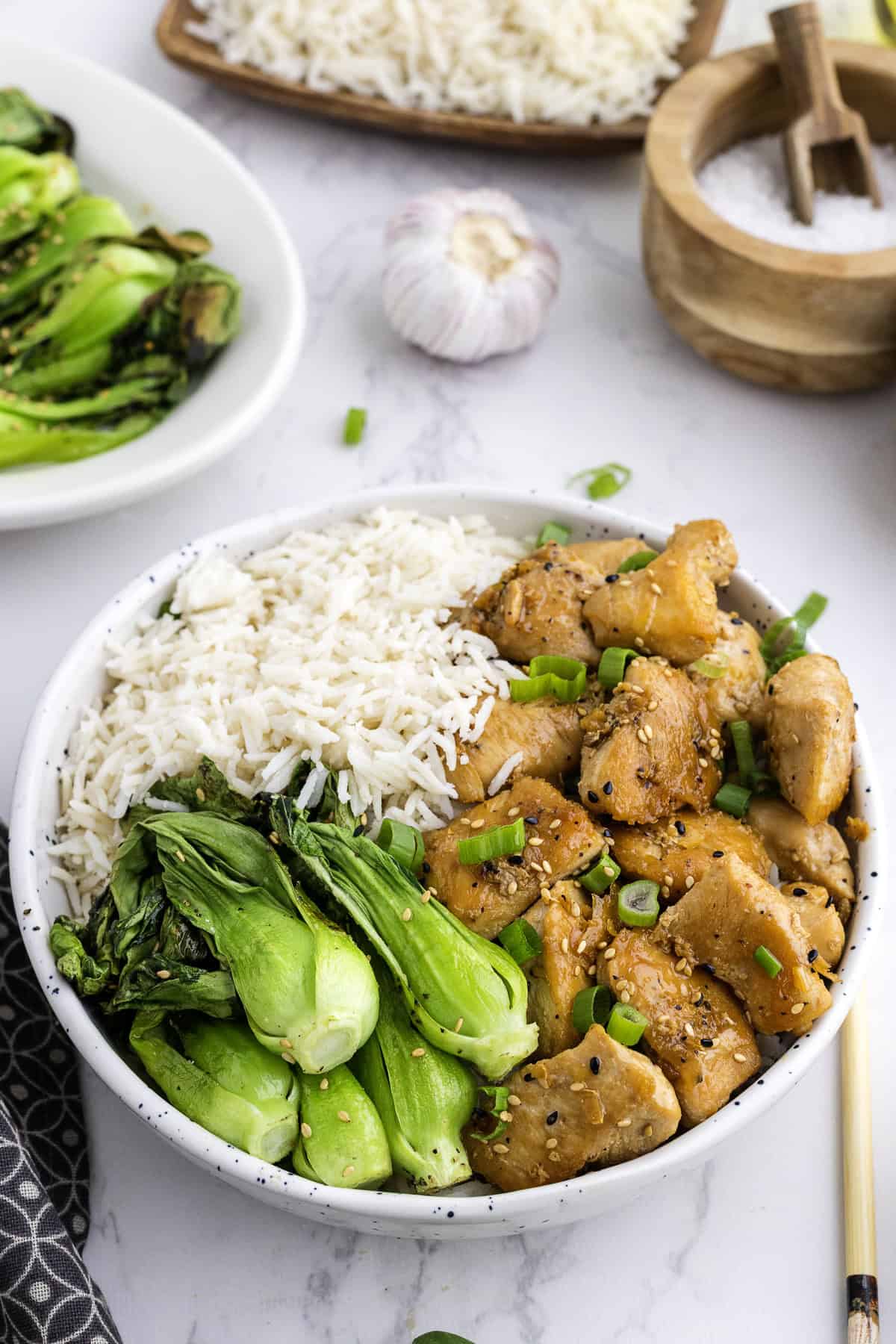 Un bol contenant du riz blanc, du bok choy cuit et des morceaux de poulet glacés garnis d'oignons verts et de graines de sésame, avec de l'ail et d'autres plats en arrière-plan.
