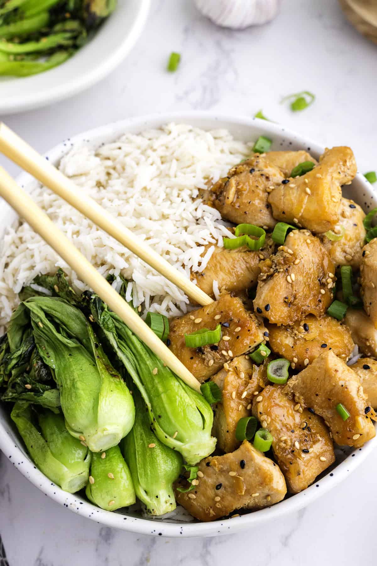 Un bol de riz blanc, du bok choy sauté et des morceaux de poulet glacés au sésame et garnis d'oignons verts, avec des baguettes posées sur le bol.