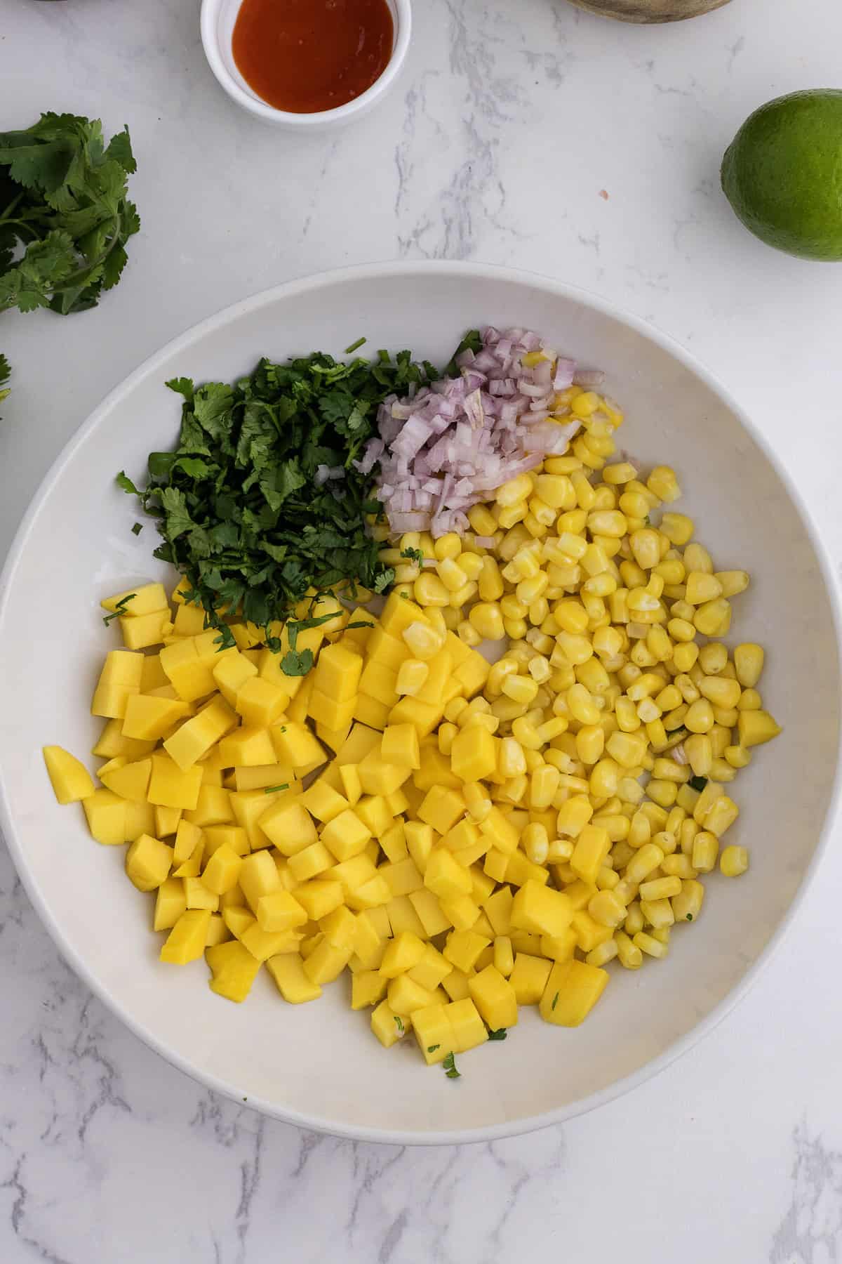 A white bowl contains chopped cilantro, diced shallot, corn, and diced mango, arranged in separate sections. A lime and a small bowl of red sauce are nearby on the marble surface.