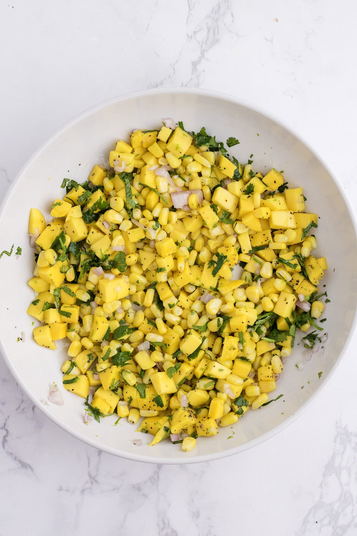 A white bowl containing a mixture of diced mango, corn, red onion, cilantro, and seasonings on a white marble surface.