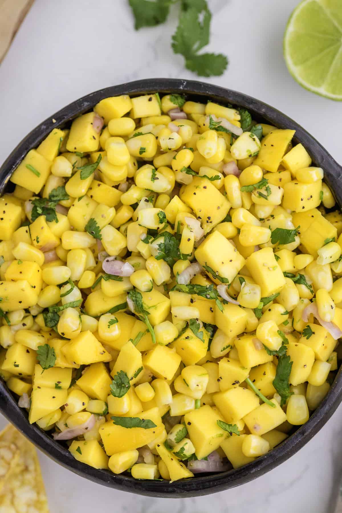 A bowl of mango corn salsa with chopped mango, corn, red onion, and cilantro, with a lime wedge and cilantro leaf on the side.