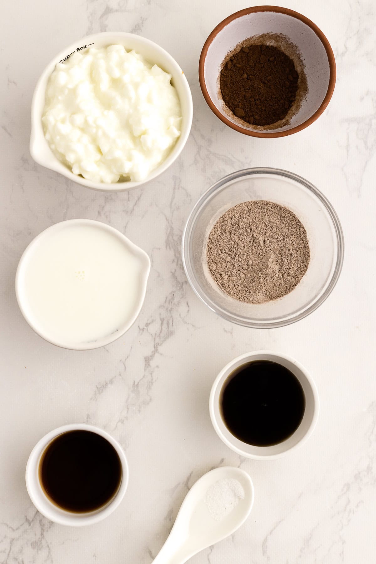 Six small bowls on a white surface containing cottage cheese, cocoa powder, milk, chocolate pudding, vanilla extract, maple syrup, and a spoonful of salt.