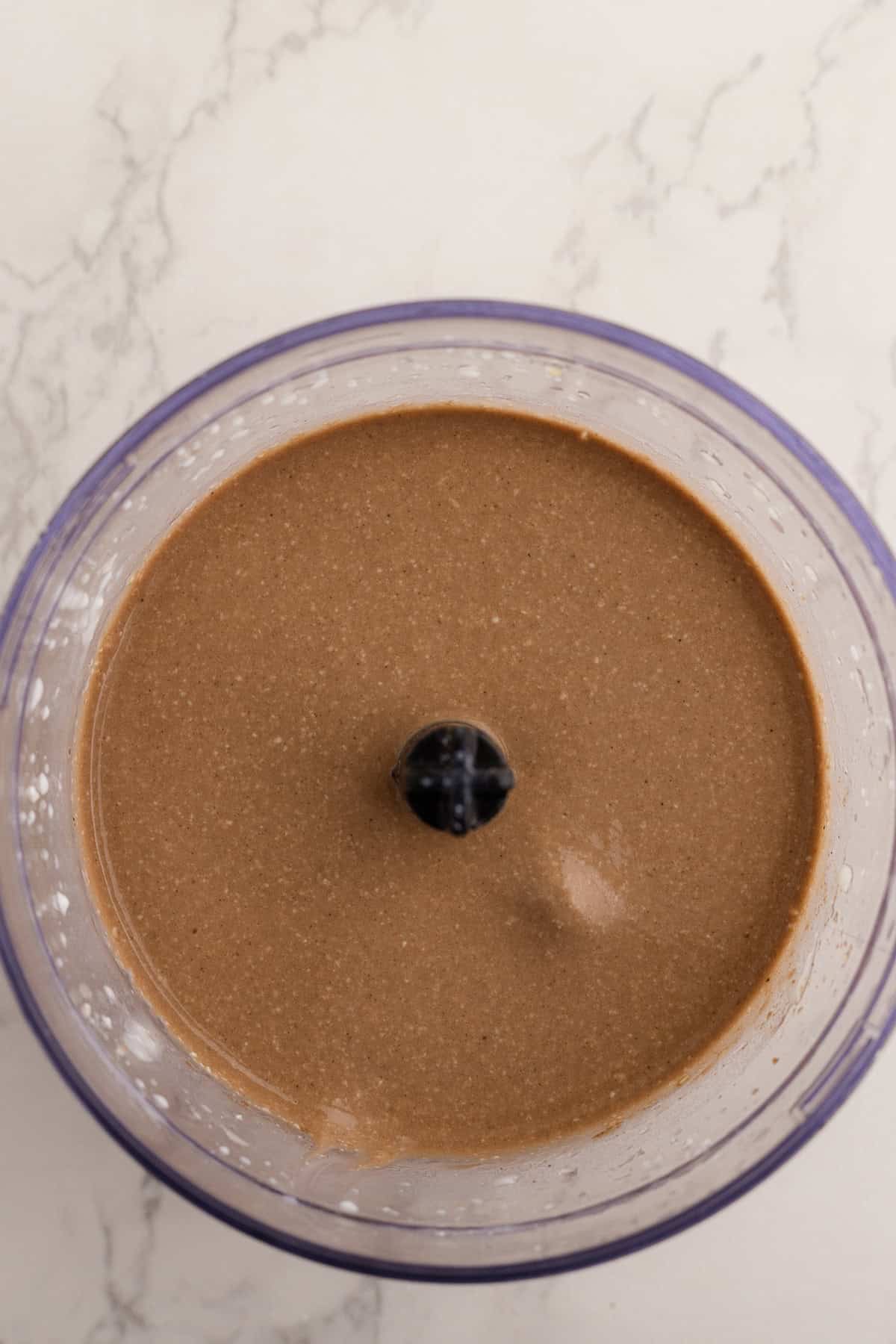 A food processor bowl filled with smooth, chocolate-colored batter sits on a light marble surface.