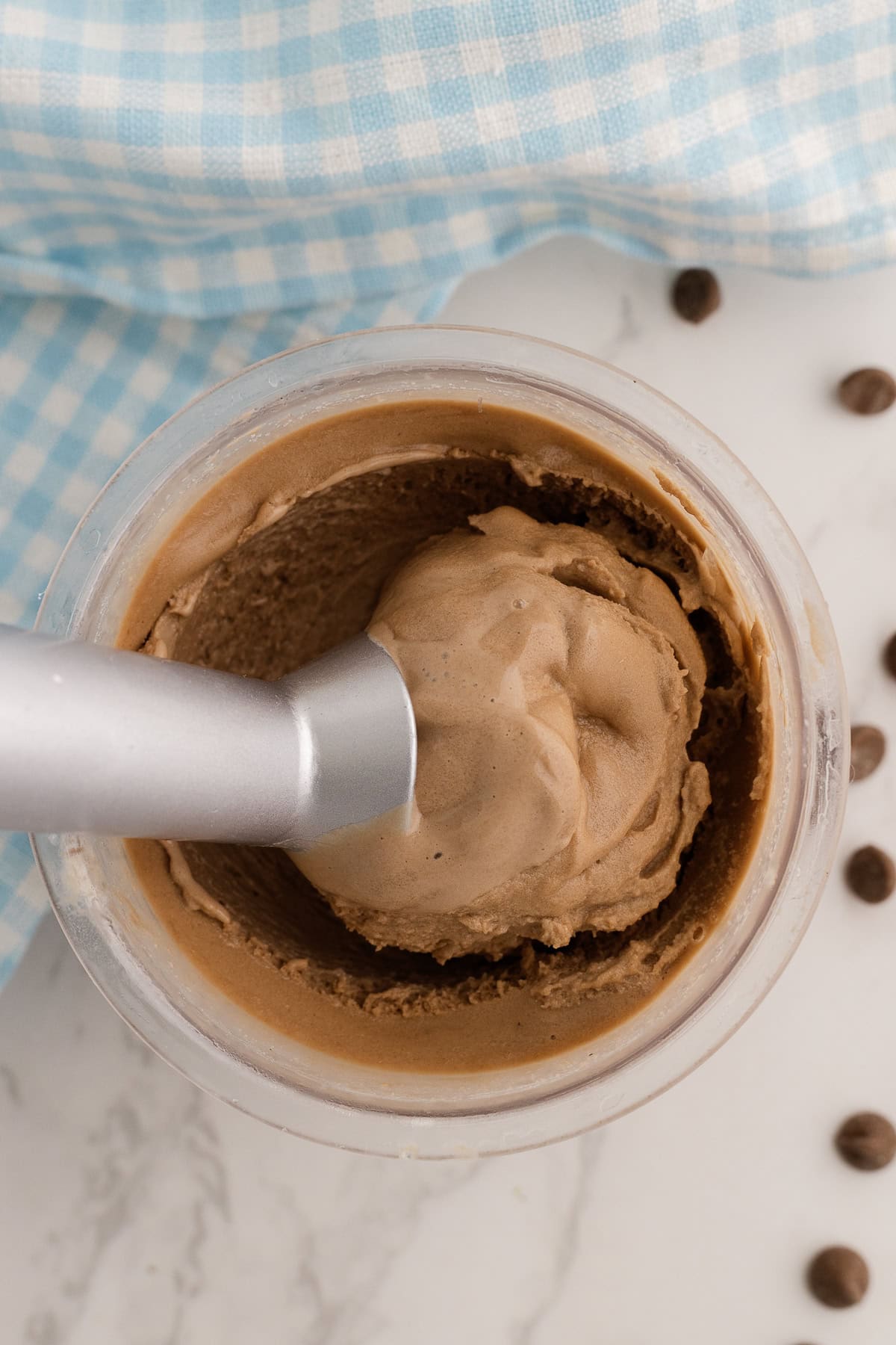 A close-up of chocolate ice cream being scooped from a container, with a checkered blue cloth and chocolate chips nearby on a marble surface.