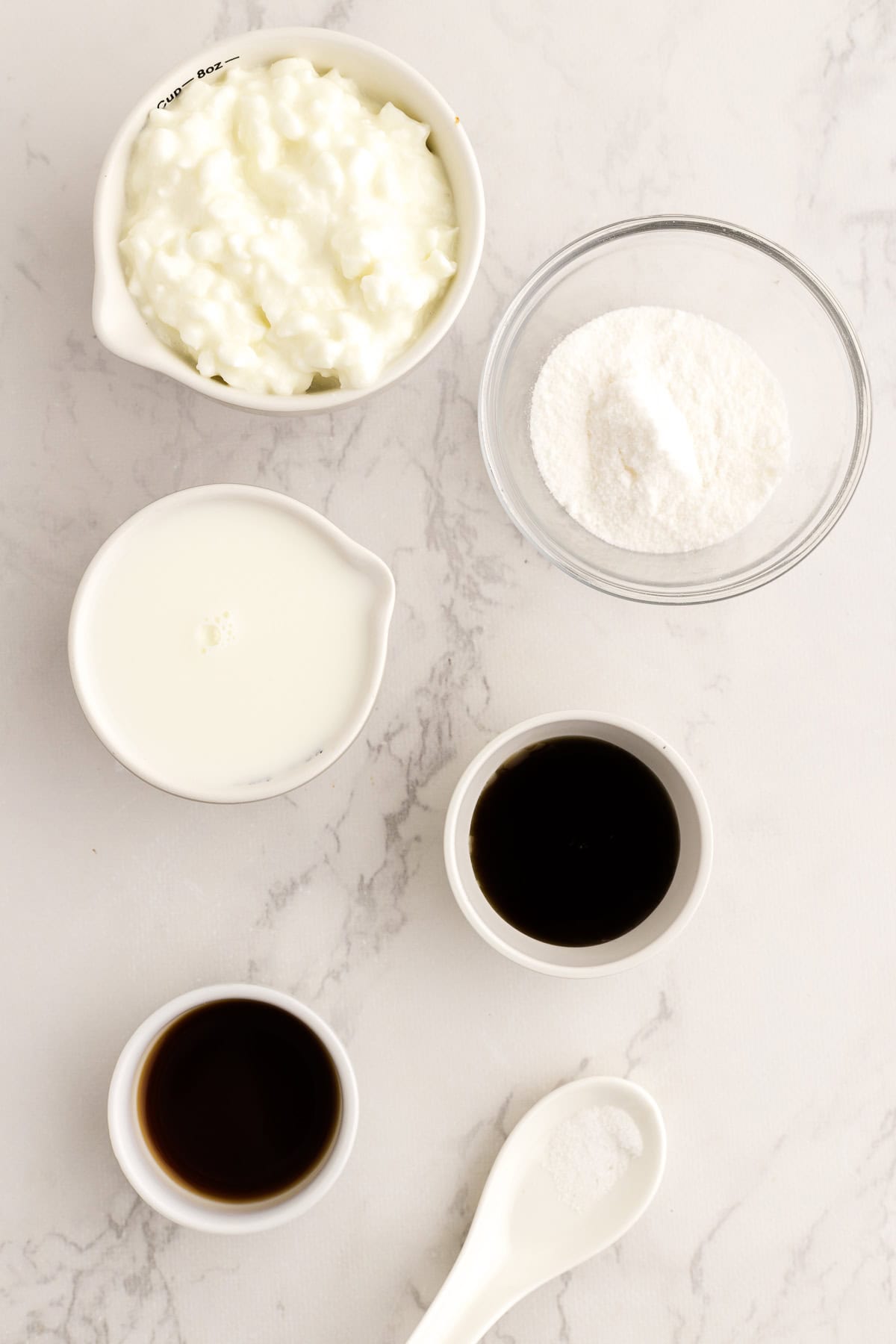 Cinq petits bols contenant du fromage blanc, du pudding a la vanolle, du lait, de l'extrait de vanille, un du sirop d'érable et une cuillère avec du sel sont disposés sur une surface claire.