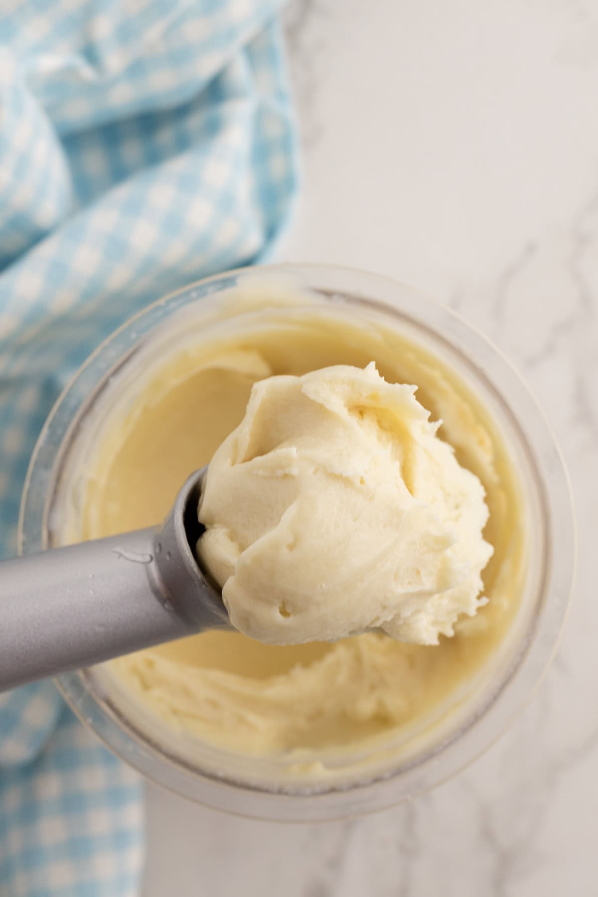 Une cuillère à glace en métal avec une boule arrondie de glace à la vanille, tenue au-dessus d'un récipient de glace, avec un tissu à carreaux bleus à l'arrière-plan.